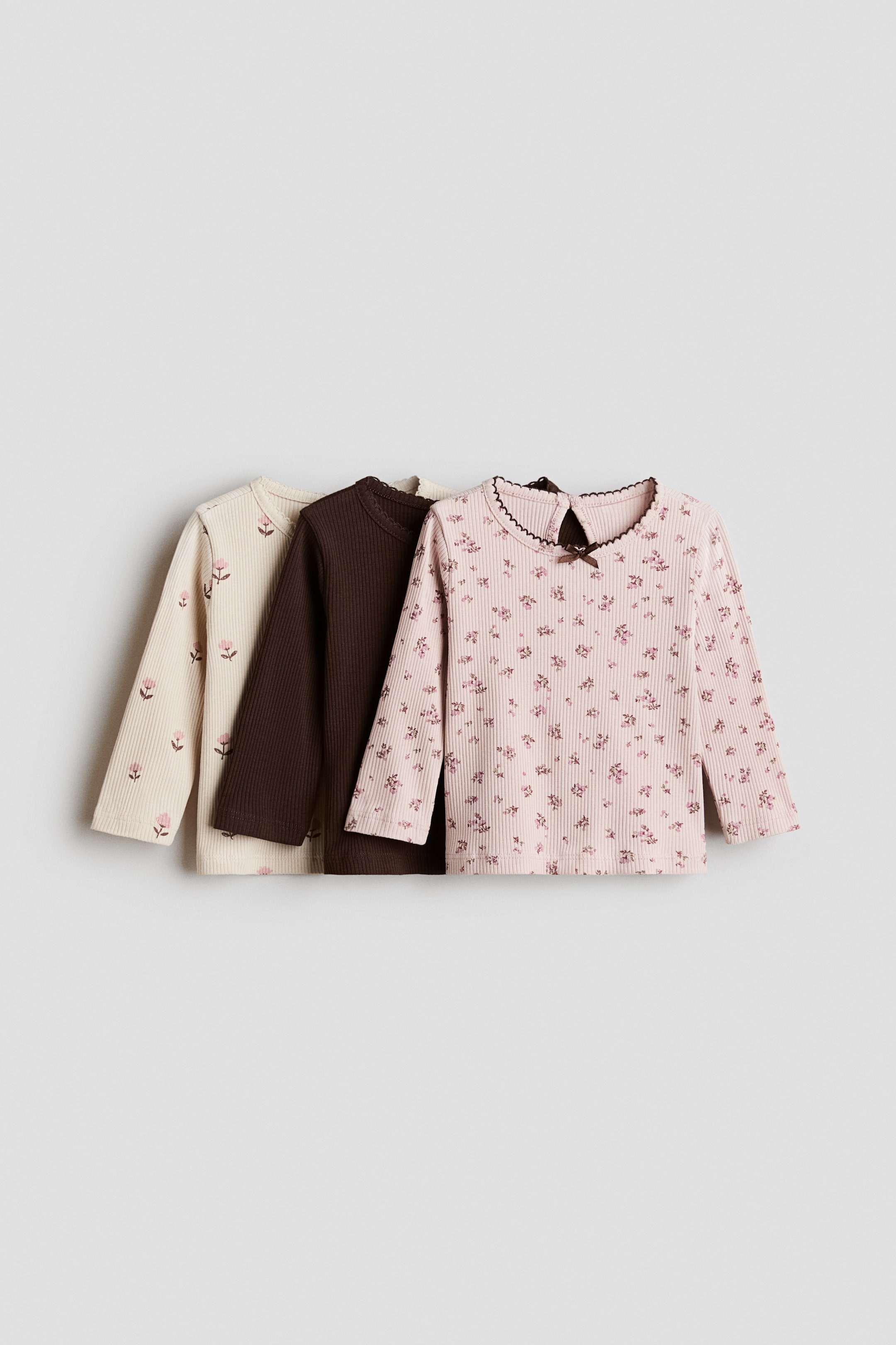 Ver imagem maior: Estão apresentados três tops de manga comprida canelados, dispostos em fila sobreposta e ligeiramente inclinados para a esquerda. O top rosa em primeiro plano apresenta um pequeno estampado floral, um decote em escalope com debrum escuro, uma abertura em gota na parte de trás do pescoço com um pequeno laço, e punhos e bainha com acabamento em ondas; atrás dele surgem um top castanho-escuro liso e um top creme com flores rosa bordadas, apresentando todos os três detalhes com acabamento em ondas.