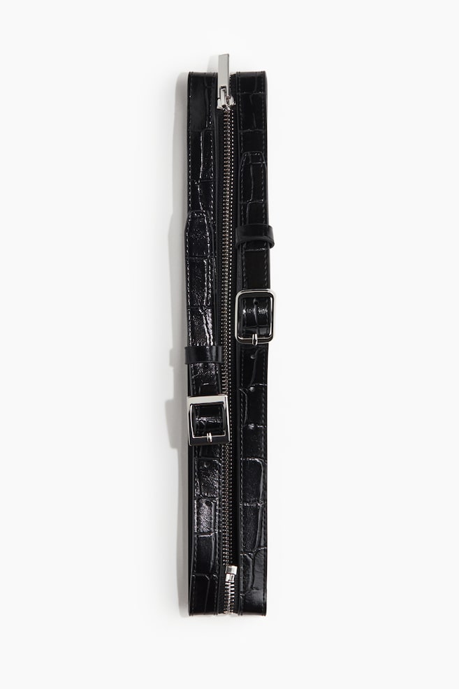 A black product is lying flat, featuring two parallel, crocodile-embossed straps. A silver zipper is running down the center between the straps, and two silver buckles are visible on the straps. - 1