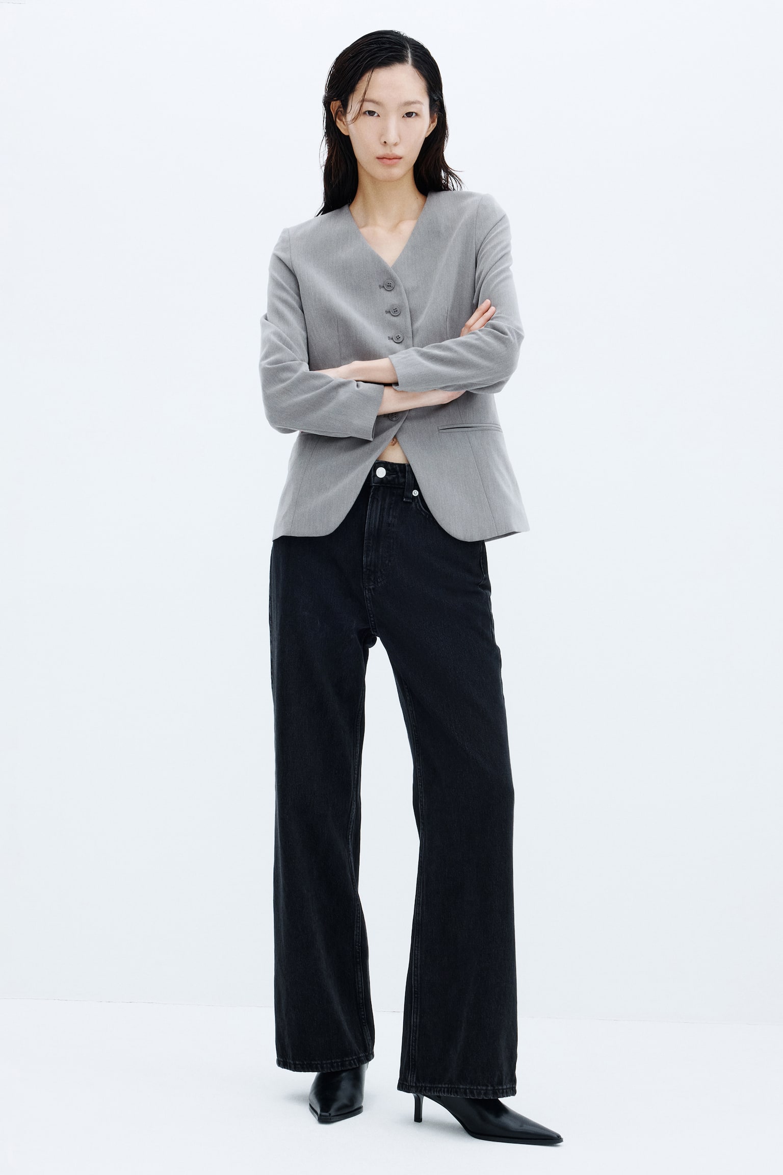 A grey lapelless twill blazer featuring a deep V-neckline, front buttons, and jetted pockets is being worn by the model standing with arms crossed. The blazer is styled with high-waisted black wide-leg jeans and black pointed-toe heeled boots. - 1