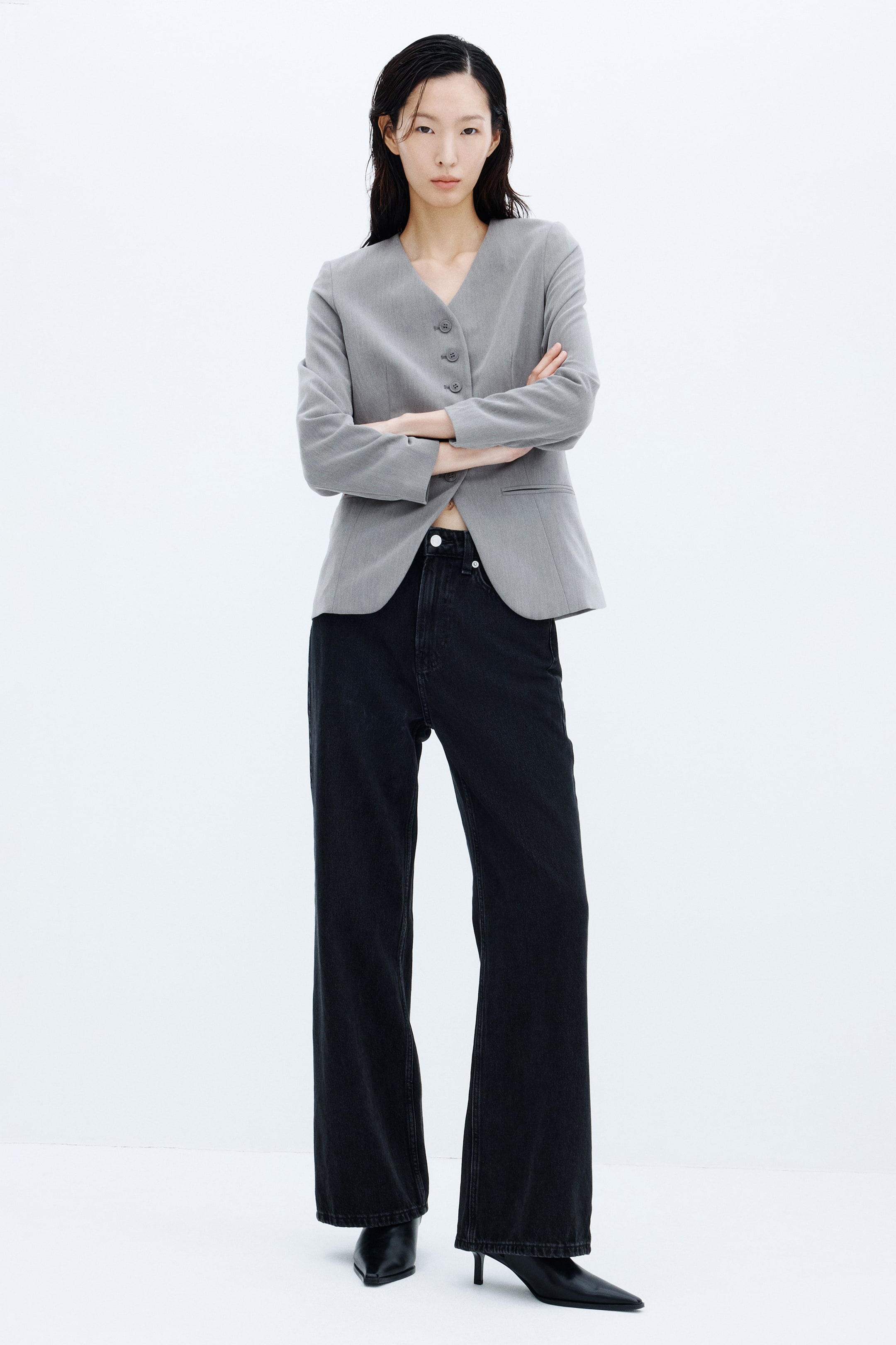 View larger image: A grey lapelless twill blazer featuring a deep V-neckline, front buttons, and jetted pockets is worn by the model, who stands with her arms crossed. The blazer is styled with high-waisted black wide-leg jeans and black pointed-toe heeled boots.