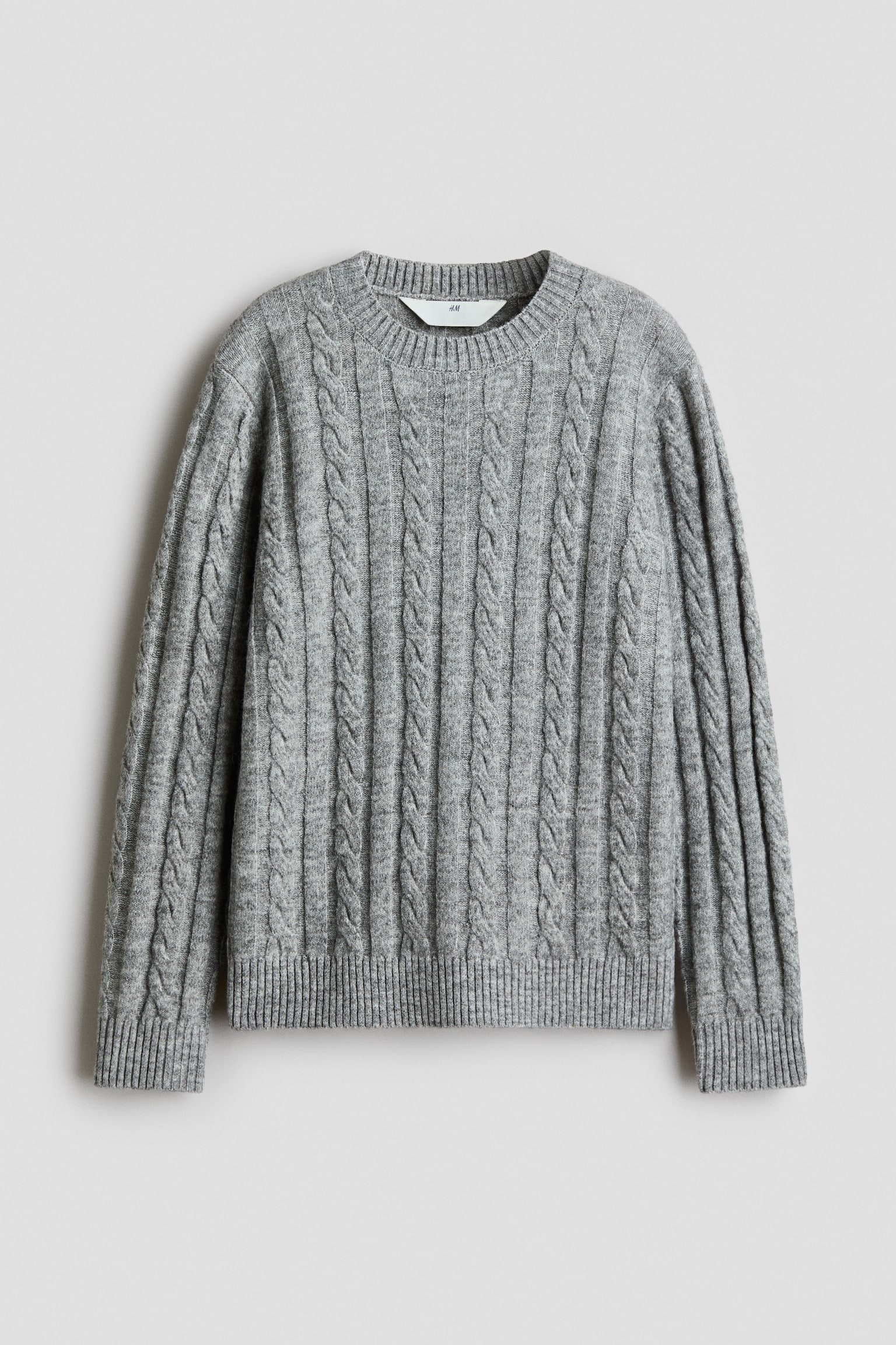 En grÃ¥ kabelstrikket sweater ses forfra og har et lodret kabelfletmÃ¸nster over hele kroppen samt lange Ã¦rmer. Den er designet med en ribstrikket rund hals, ribstrikkede manchetter og en matchende ribstrikket kant.