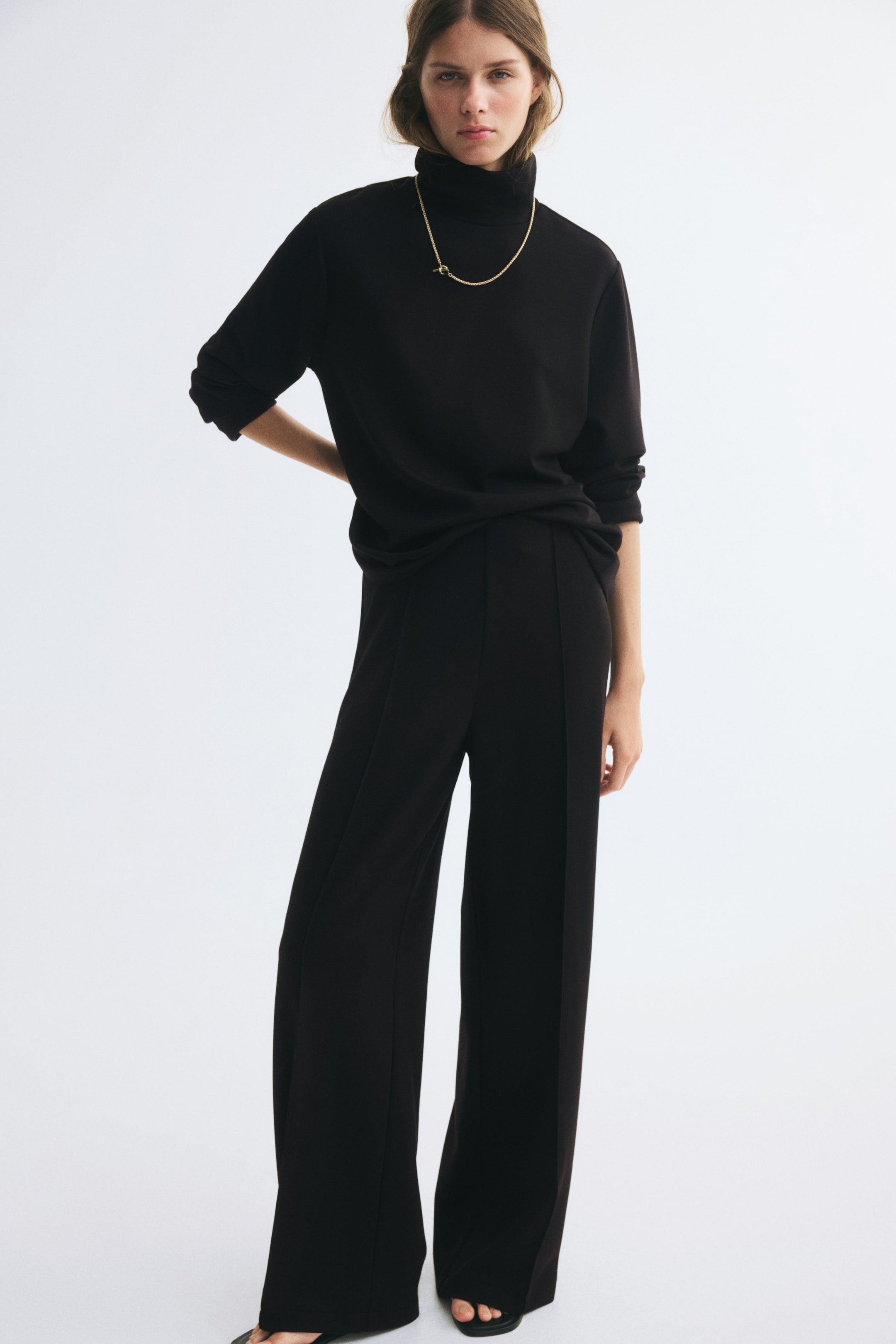 View larger image: A black jersey set featuring a long-sleeved top with a stand collar and high-waisted, straight-leg trousers with front creases is worn by the model. The top is styled with a simple gold chain necklace as the model stands facing forward.