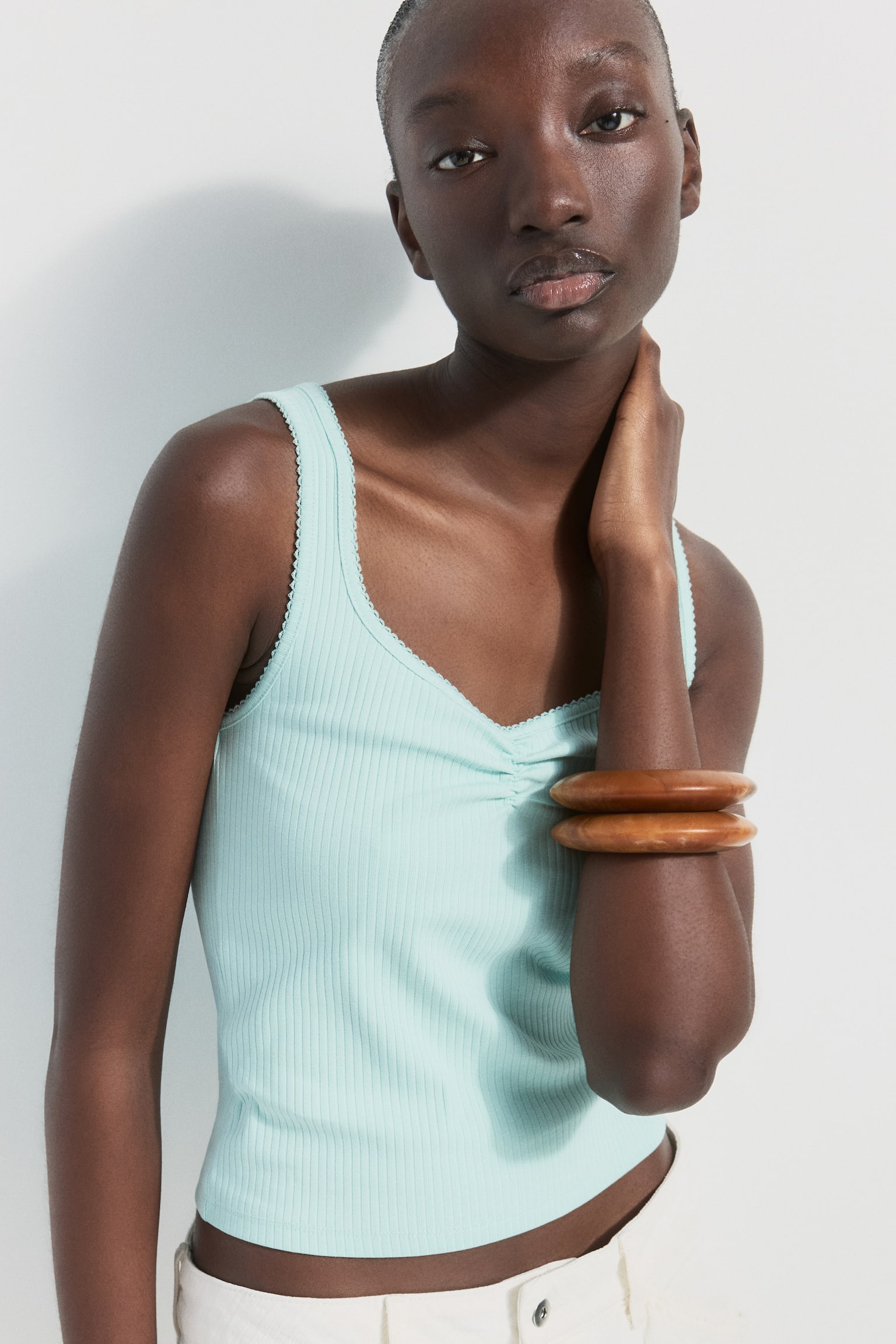 A light blue fitted vest top in ribbed cotton jersey features a sweetheart neckline with a ruched seam at the front and delicate picot trim around the neckline and armholes. The model is wearing the top, with one hand touching their neck, and two wooden bangles are visible on their forearm. - 1
