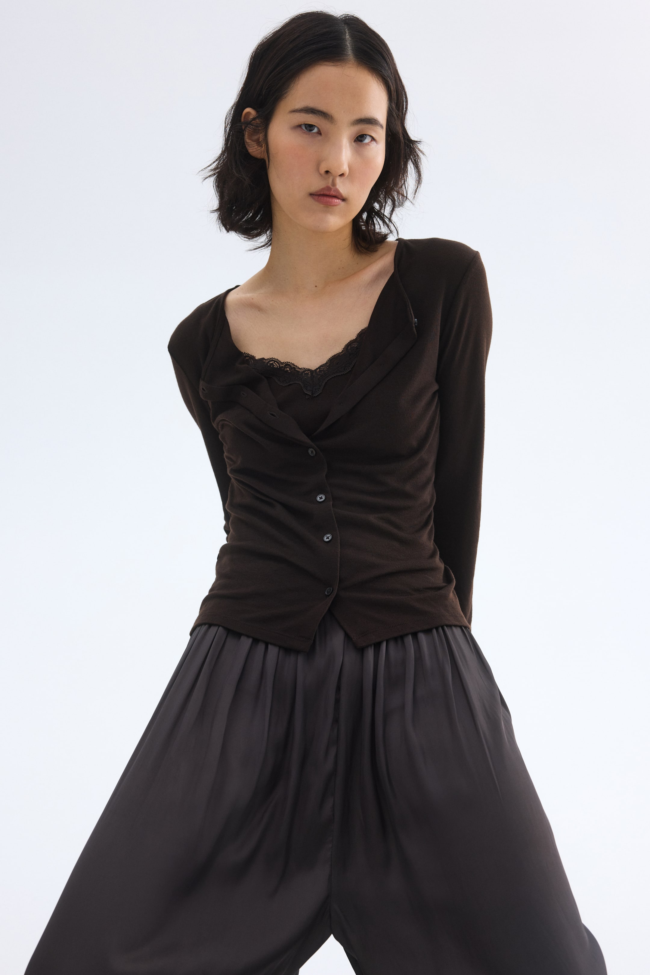 Vis større bilde: En mørkebrun langermet topp med delvis kneppet front, bred drapert halslinje og subtil rynking, lagvis over en mørkebrun camisole med svart blondekantet V-hals. Toppen og camisolen kombineres med mørkegrå bukser med vide ben, som fremhever folder i midjen og en myk glans, alt presentert fra en frontvendt, lett vinklet perspektiv.