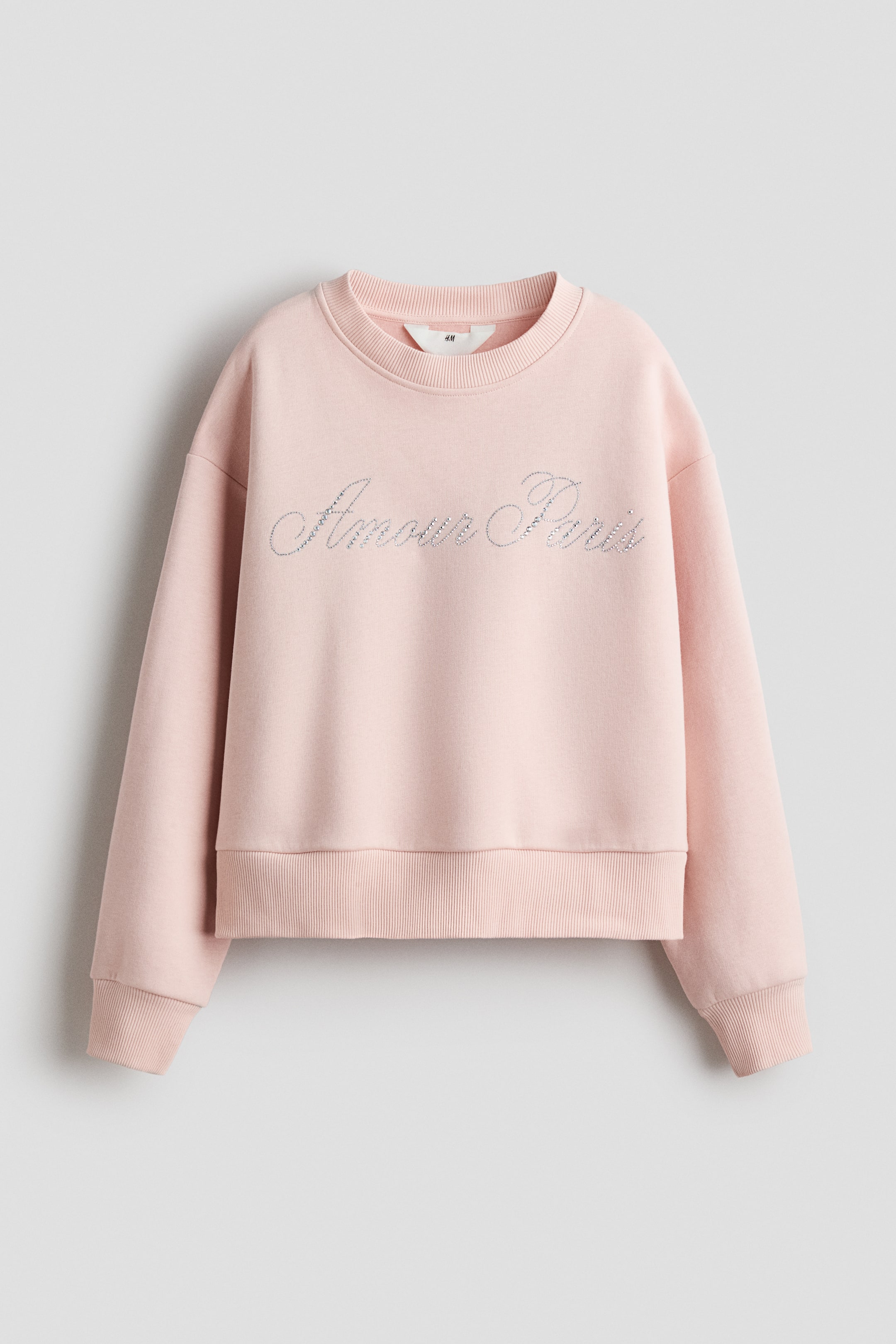 Ver imagen más grande: Una sudadera de color rosa claro muestra su PARTE DELANTERA, con un motivo reluciente de "Amour Paris". Está diseñada con un cuello redondo acanalado, hombros caídos, y puños y dobladillo acanalados.