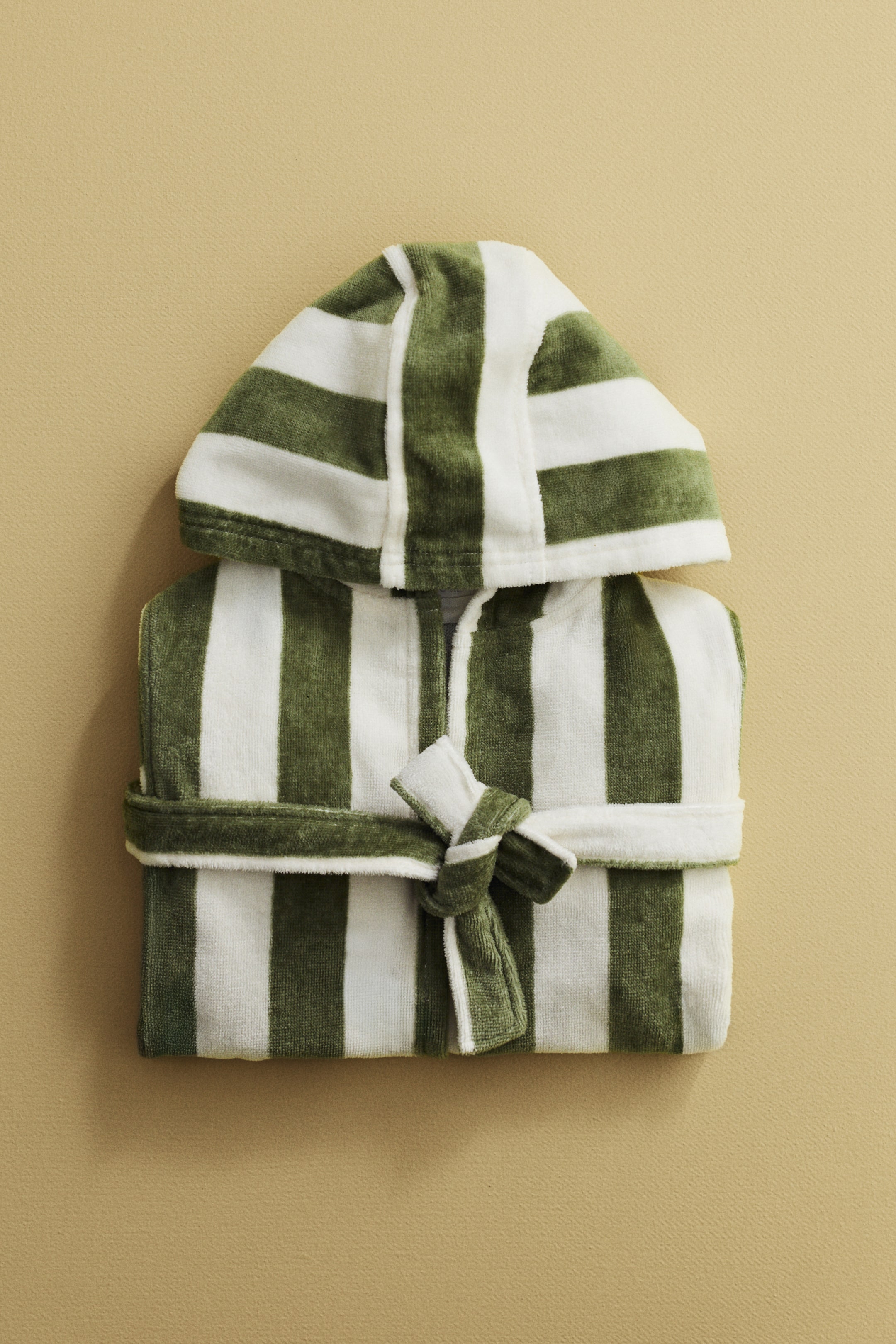 Nagyobb kép megtekintése: Egy zöld-fehér csíkos frottír köntös fekszik kiterítve, kapucnival és a középen megkötött, hozzáillő övvel. A köntös hangsúlyos függőleges csíkokat mutat be, váltakozó zöld és fehér színekben.