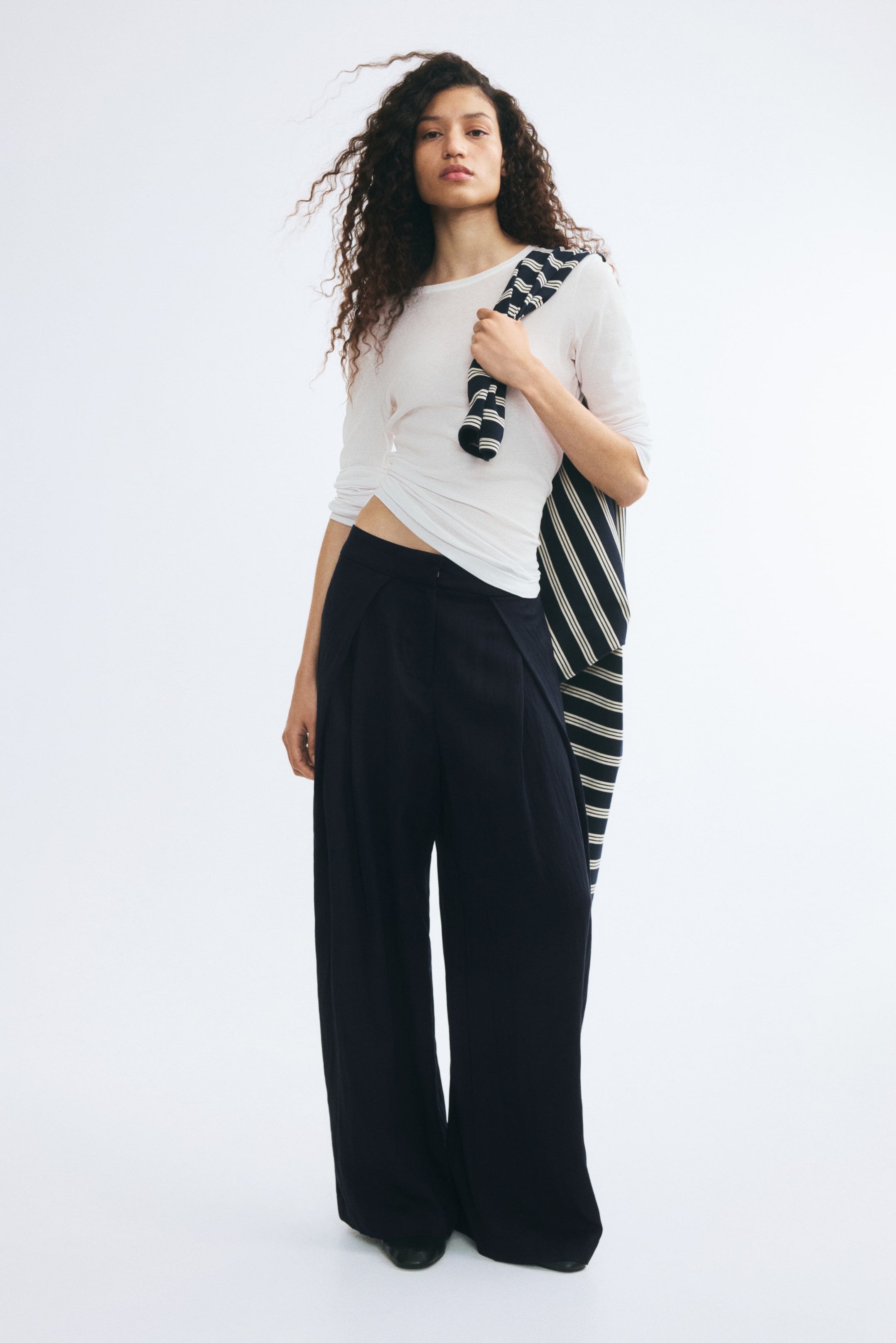 Agrandir l'image: Pantalon barrel noir à coupe ample, doté de plis sur le devant et d’un détail portefeuille distinctif au niveau des hanches, porté par le mannequin. Le pantalon est assorti à un haut blanc à manches longues et à un pull en maille rayé drapé sur une épaule.