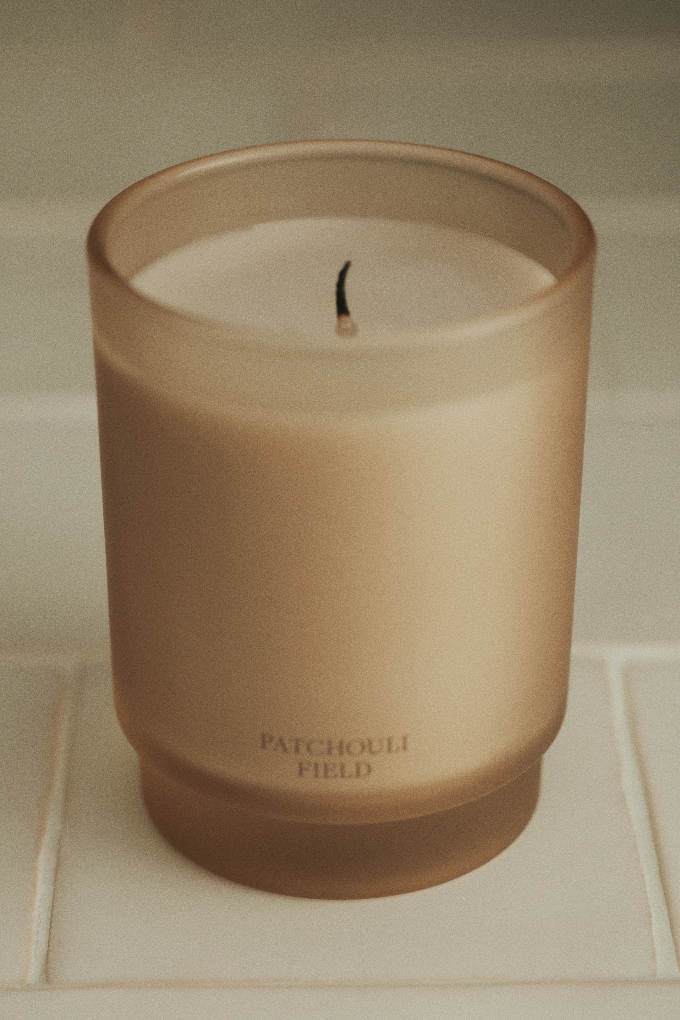 En rektangulær produktæske står oprejst, og viser sin forside og venstre side. Forsiden viser "PATCHOULI FIELD" branding og produktdetaljer, mens den synlige side lister duftnoter. Til højre står et matteret glaslys, der indeholder lysfarvet voks og har "PATCHOULI FIELD" på forsiden.