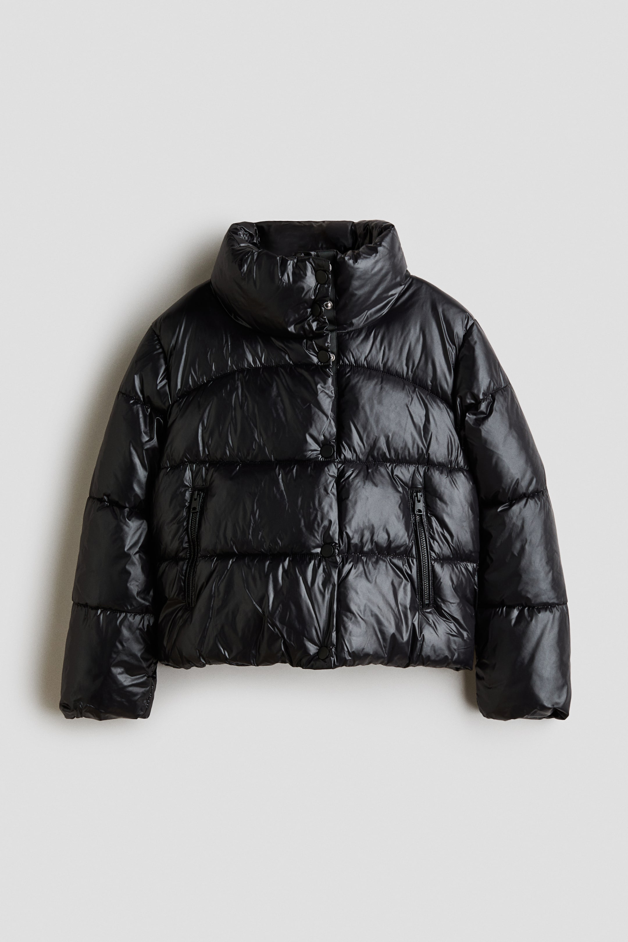 Ver imagen más grande: Se muestra una chaqueta puffer acolchada corta en un tejido acolchado negro brillante, vista de frente, con cuello alto y pespuntes horizontales. La chaqueta presenta cierre frontal con botones a presión, bolsillos verticales con cremallera y mangas largas con puños elásticos.