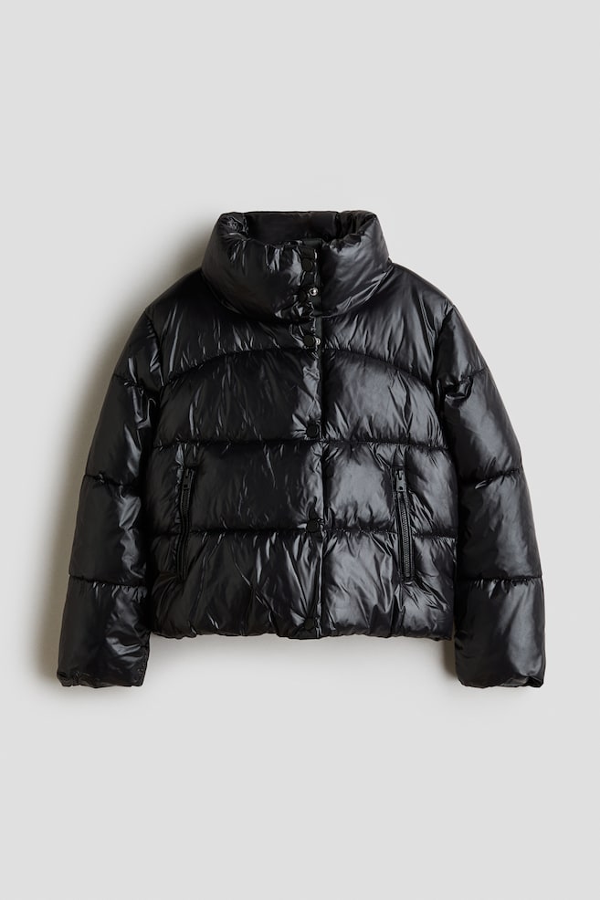 A short, padded puffer jacket in a shiny black quilted fabric is shown facing forwards, featuring a high stand collar and horizontal quilting. The jacket features a snap-button front fastening, vertical zip pockets, and long sleeves with elasticated cuffs. - 1