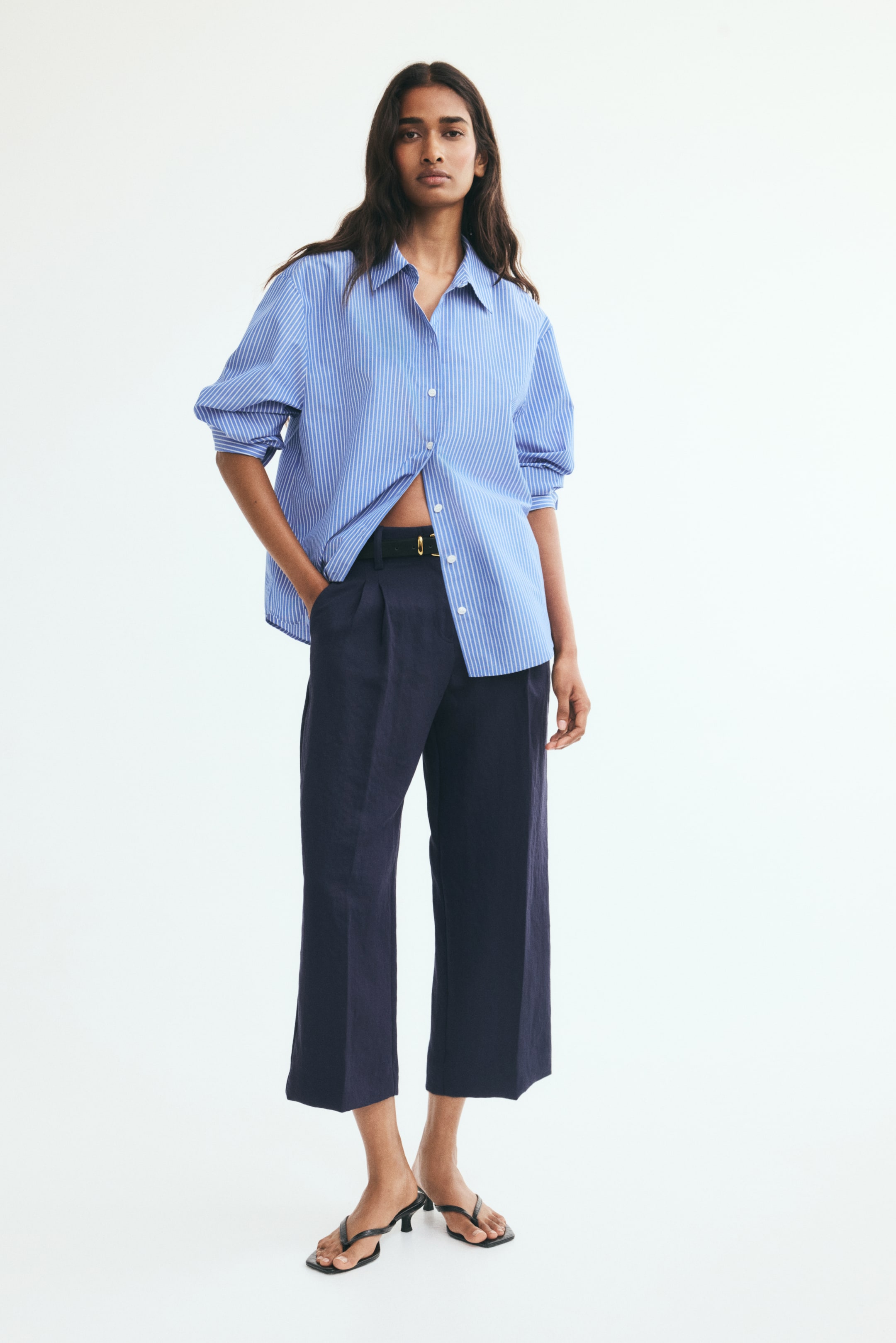 View larger image: Tailored navy blue ankle-length pants with front pleats and straight legs are being worn by the model, who is resting one hand inside the diagonal side pocket. The pants are styled with a black belt, an oversized blue and white striped button-up shirt, and black heeled sandals.