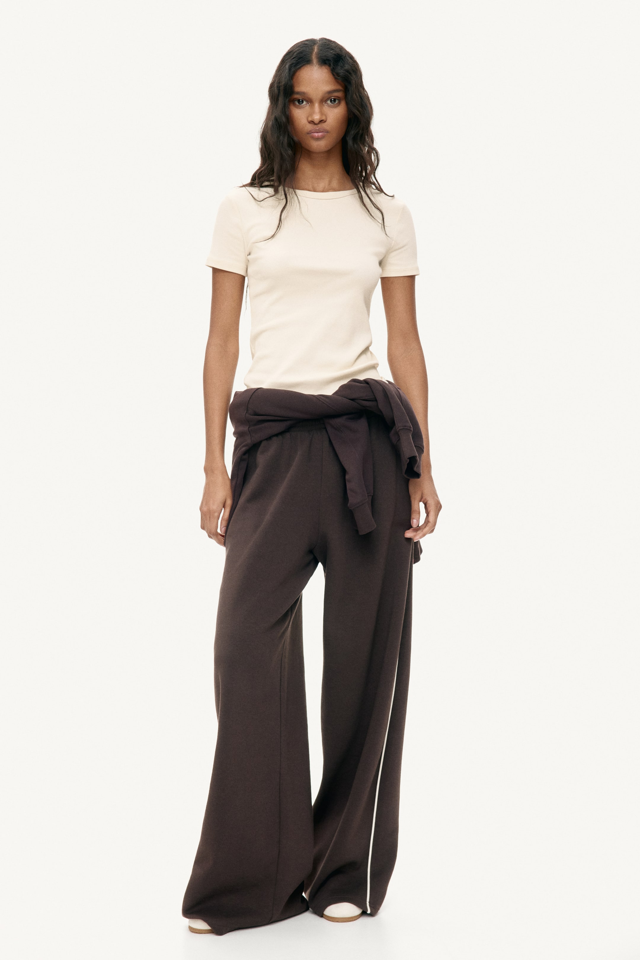 הצגת תמונה גדולה יותר: חולצת טי שירט בצבע קרם עם שרוולים קצרים ומרקם מצולע, מאופיינת בצווארון עגול וגזרה צמודה. מכנסיים רחבים בצבע חום כהה מציגים פס לבן לאורך התפר הצדדי הימני, וסווטשירט תואם בצבע חום כהה מעוצב קשור סביב המותניים.