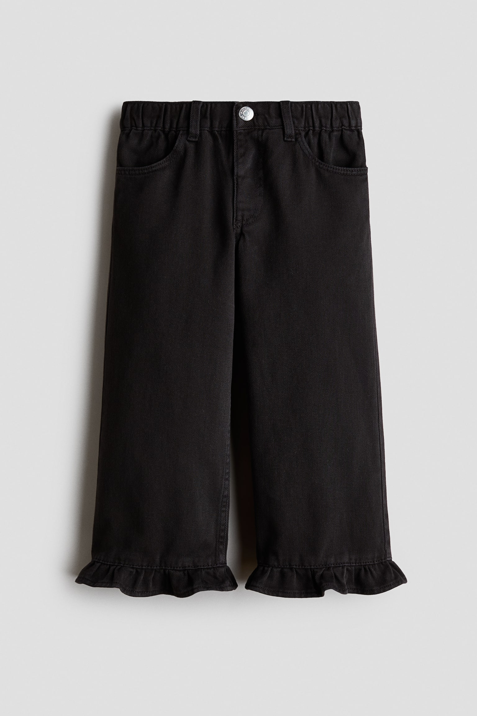 Un pantalon en denim noir est prÃ©sentÃ© de face, avec une taille Ã©lastiquÃ©e munie de passants pour ceinture, des poches avant arrondies et une fermeture par un seul bouton argentÃ©. Le pantalon prÃ©sente une coupe droite qui se termine par des ourlets volantÃ©s en bas.