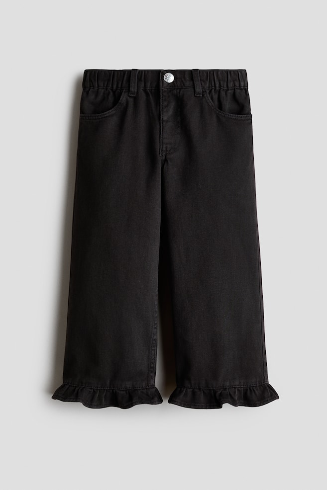 Ett par svarta denimbyxor visas framifrÃ¥n, med en resÃ¥rmidja med bÃ¤lteshÃ¤llor, rundade framfickor och en enkel knappknÃ¤ppning i silverfÃ¤rg. Byxorna har en rak benvidd som avslutas med volangkant nedtill. - 1