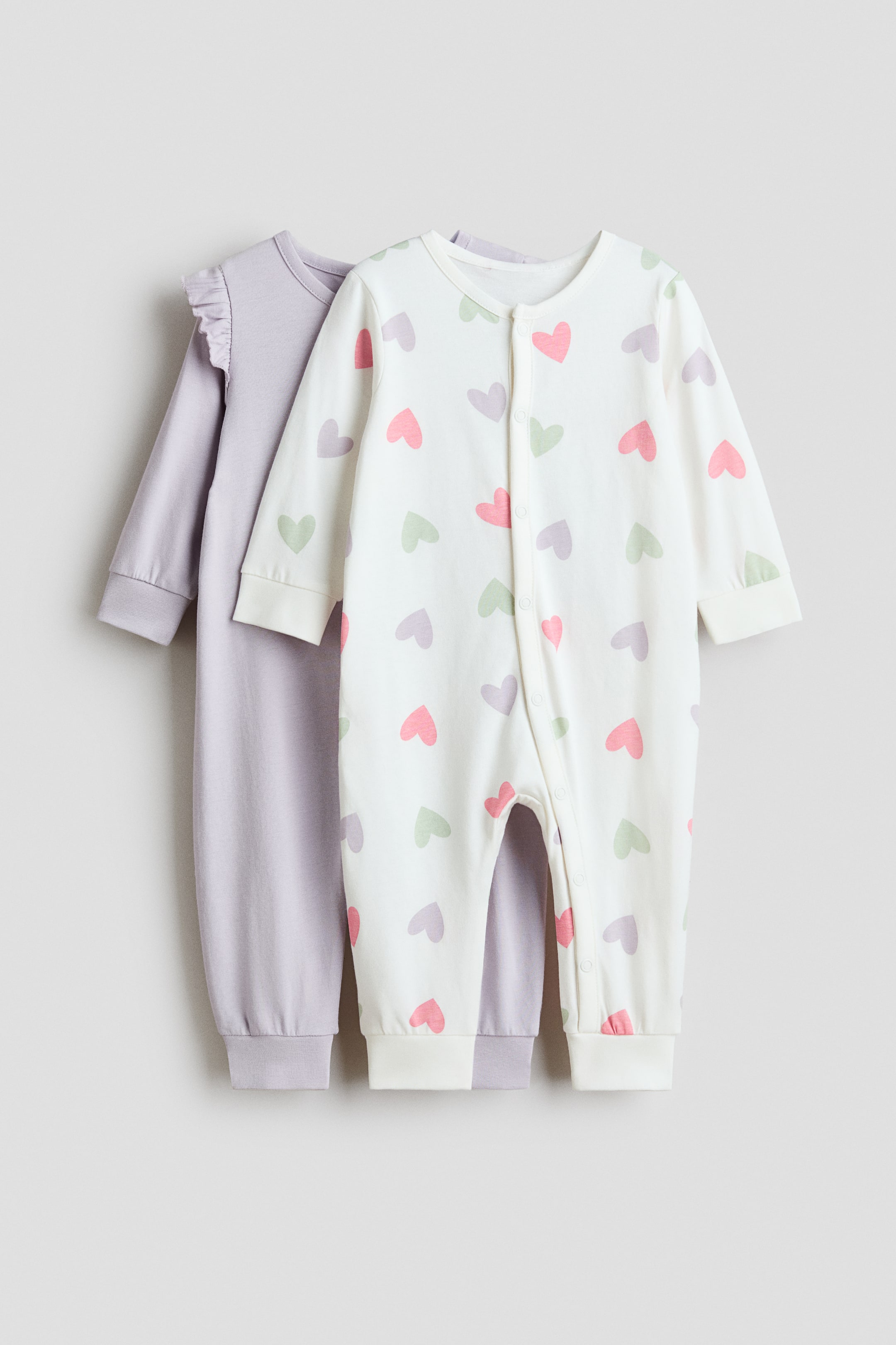 Agrandir l'image: Deux pyjamas tout-en-un se tiennent debout, avec le pyjama blanc à l'avant arborant un motif de cœurs roses, vert clair et violet clair. Ce pyjama est conçu avec des manches longues, des ourlets resserrés aux poignets et aux chevilles, et des boutons-pression qui descendent le long du devant et d'une jambe. Il est superposé devant un pyjama violet clair uni qui présente des détails froncés aux épaules et des ourlets resserrés.