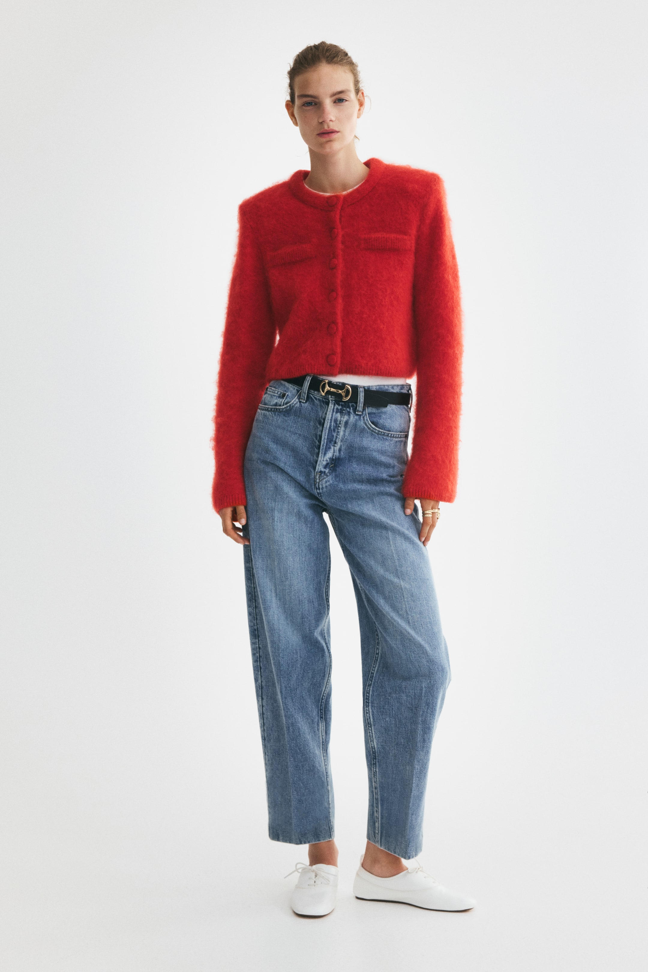 Größeres Bild anzeigen: Das Model trägt eine kurze, rote Cardigan aus weichem Strick über einem weißen Unterhemd und mit blauen Jeans. Sie ist mit einem runden Halsausschnitt gestaltet, verdeckte Knöpfe verschließen die Vorderseite, und dekorative Brusttaschen sind auf der Brust sichtbar; Schulterpolster sorgen für eine definierte Silhouette, und schmale Rippenabschlüsse vollenden die Ärmelbündchen und den Saum.