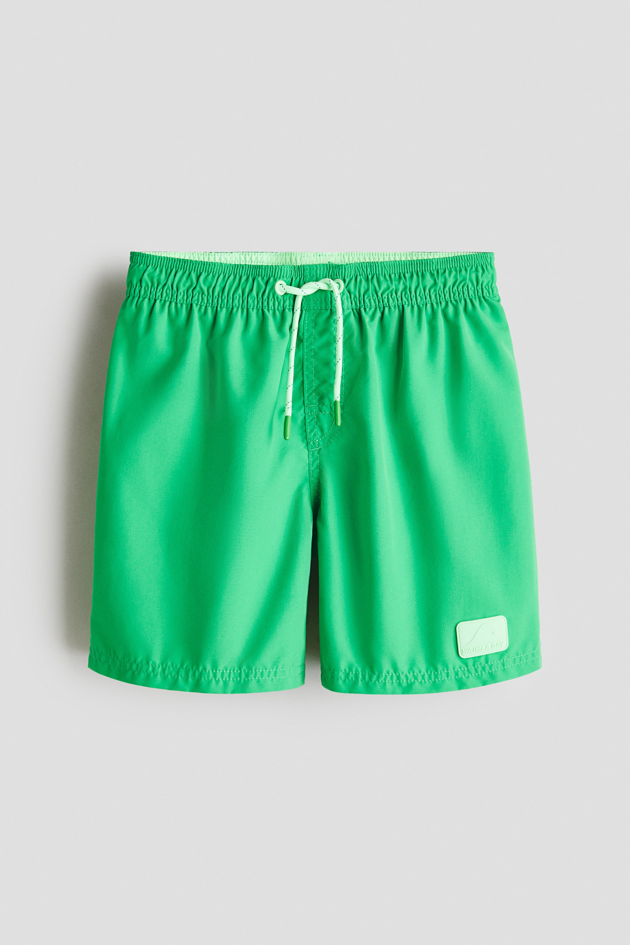 Größeres Bild ansehen: Grüne Badeshorts in Vorderansicht, mit einem elastischen Bund, einer weißen Kordel mit grünen Enden und einem hellgrünen rechteckigen Logo-Patch auf dem unteren rechten Bein.