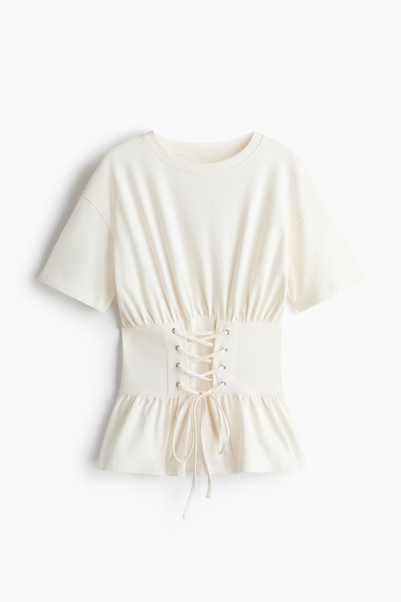 Een gebroken wit T-shirt wordt frontaal getoond, met een ronde hals, korte mouwen en een brede, aansluitende tailleband in corsetstijl met zilverkleurige oogjes en een veterlint. De stof plooit zacht boven de tailleband en loopt onderaan uit in een gerimpelde peplumzoom.