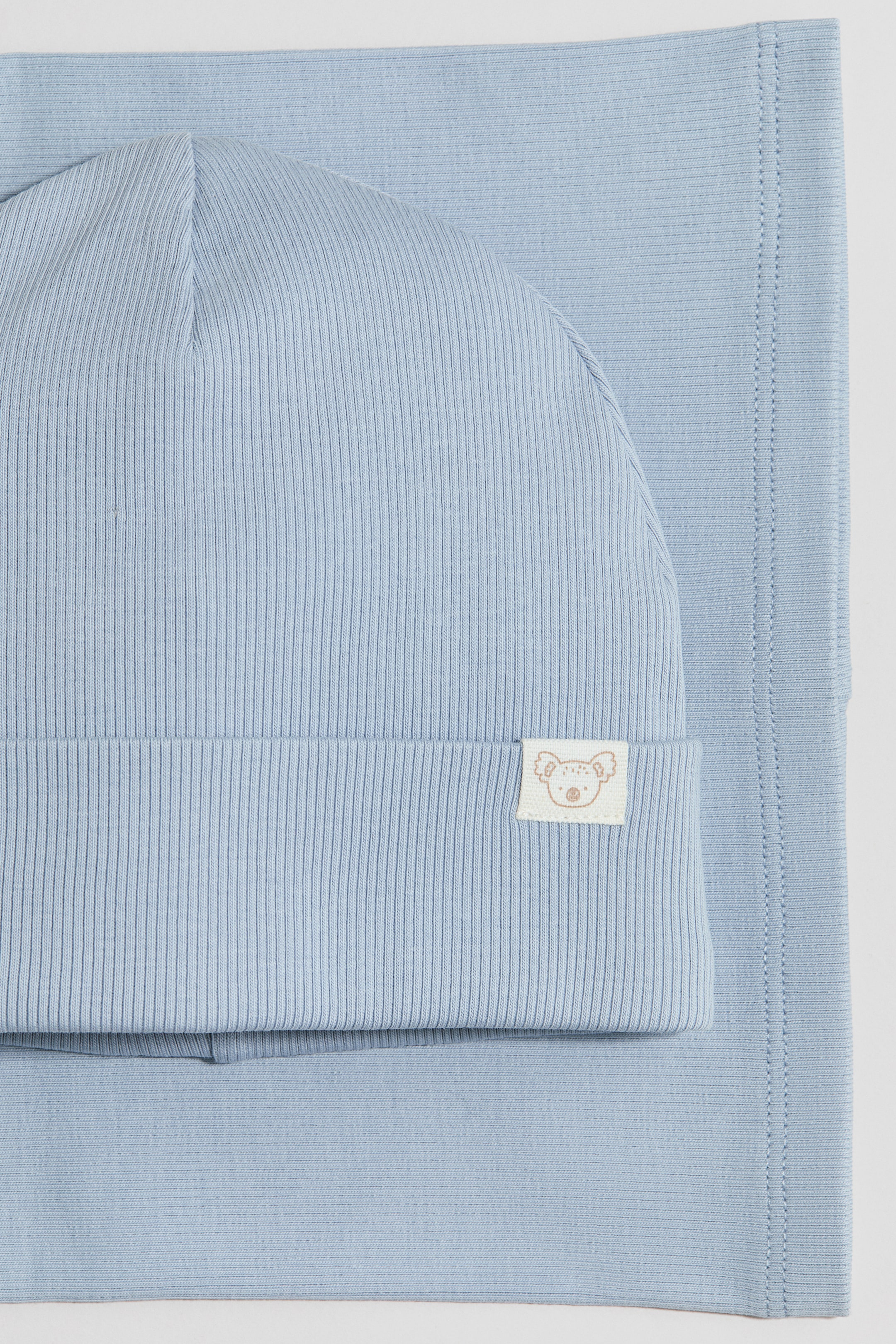 Grösseres Bild ansehen: Das hellblaue 2-teilige Jersey-Set wird präsentiert und besteht aus einer gerippten Beanie-Mütze sowie einem passenden flachen Jersey-Stoffstück. Die Beanie weist einen umgeschlagenen Bund mit einem kleinen Stoffetikett mit Koala-Motiv auf, während das flache Teil eine abgestepptte Kante zeigt.