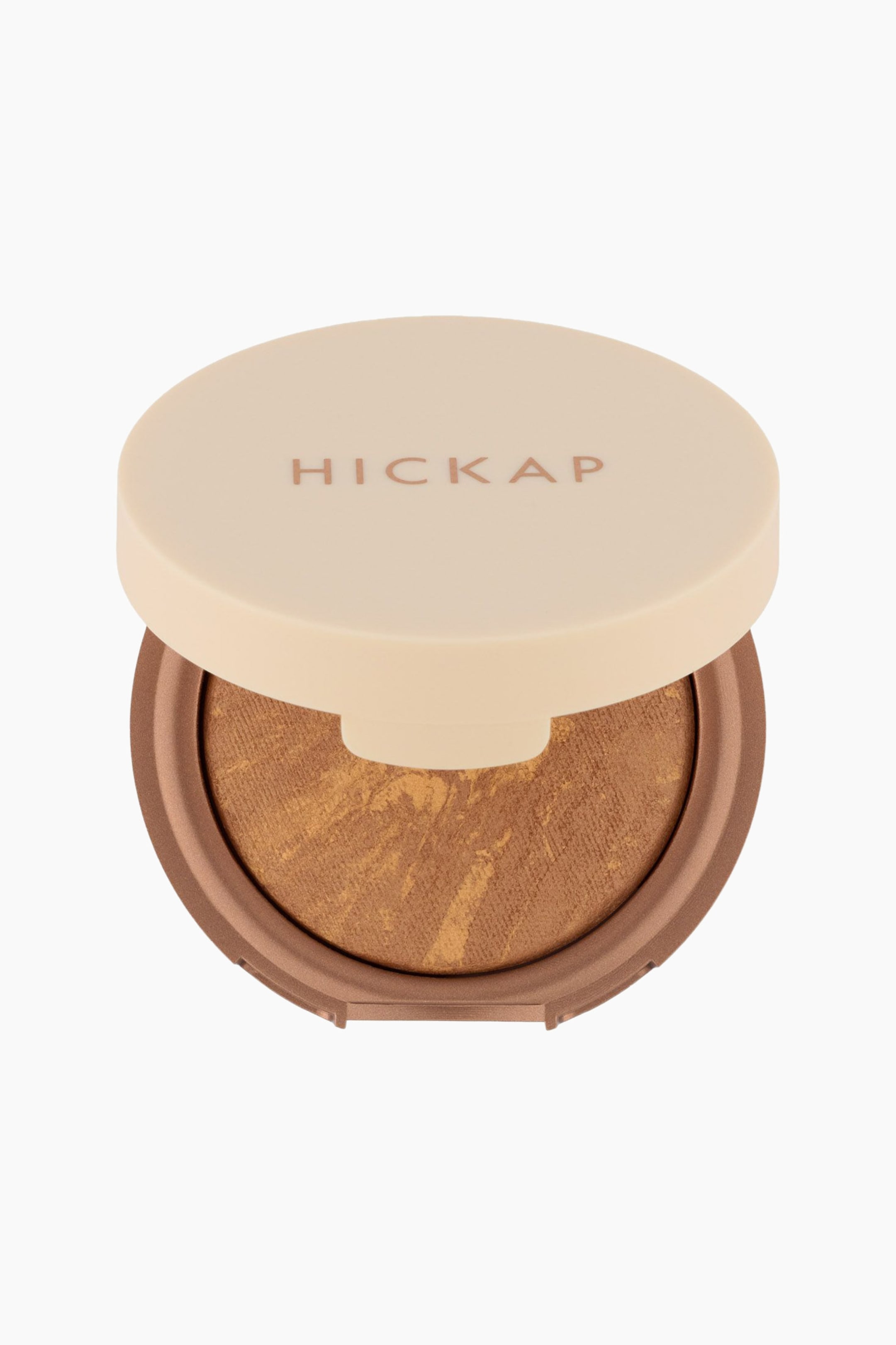 Se større billede: Et makeup-kompakt ligger let opad med låget delvist åbent, hvilket afslører produktet indeni. Det runde, cremefarvede låg har brandnavnet "HICKAP" i brune bogstaver, og kompaktets brune bund indeholder et skinnende, marmoreret bronzepulver med lysere guldstriber.