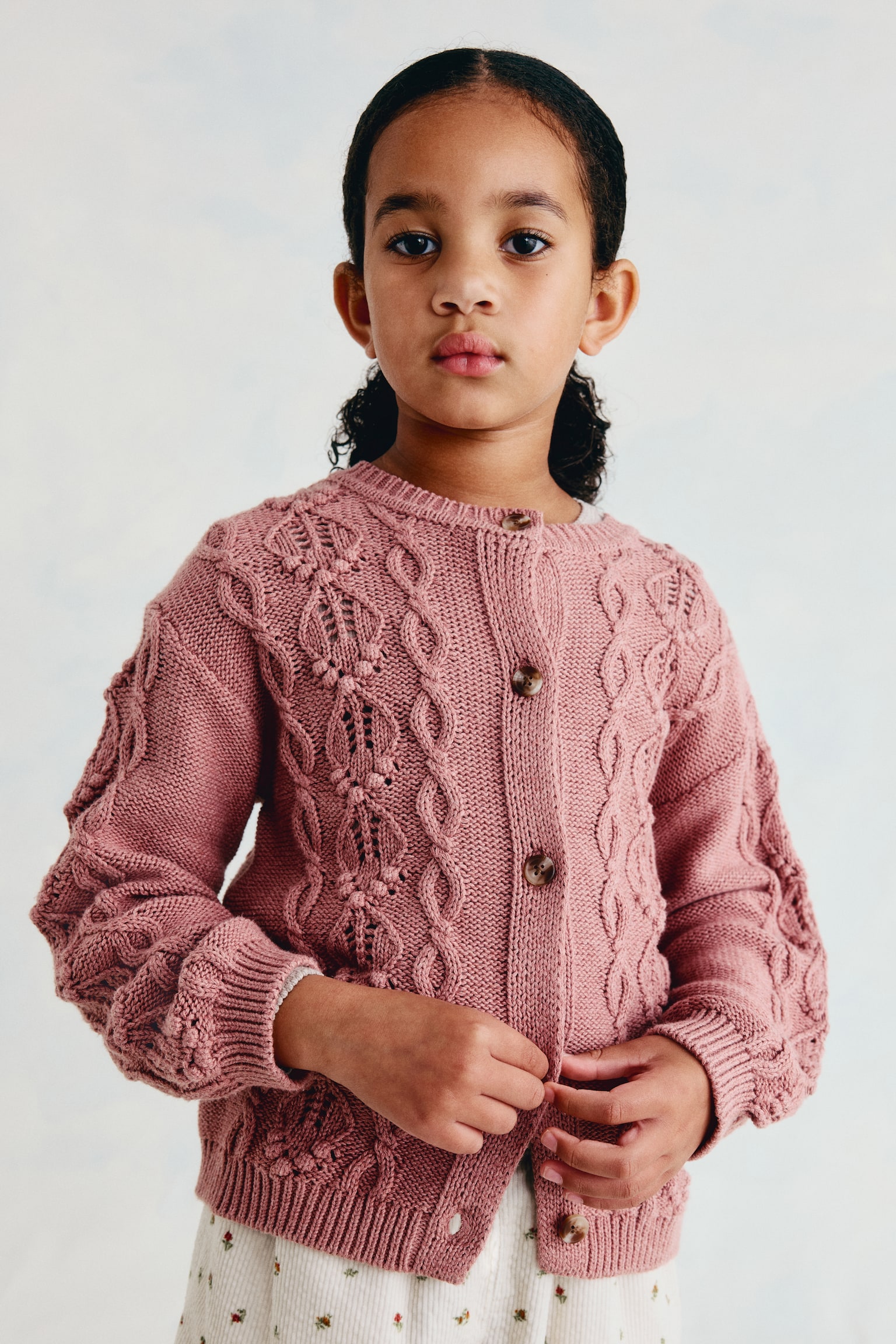 A dusty pink knitted cardigan is shown from the front, featuring intricate cable-knit and pointelle-knit patterns. It is designed with a round neckline, a button-down front with five brown buttons, dropped shoulders, long sleeves, and ribbed trim at the collar, cuffs, and hem. - 1