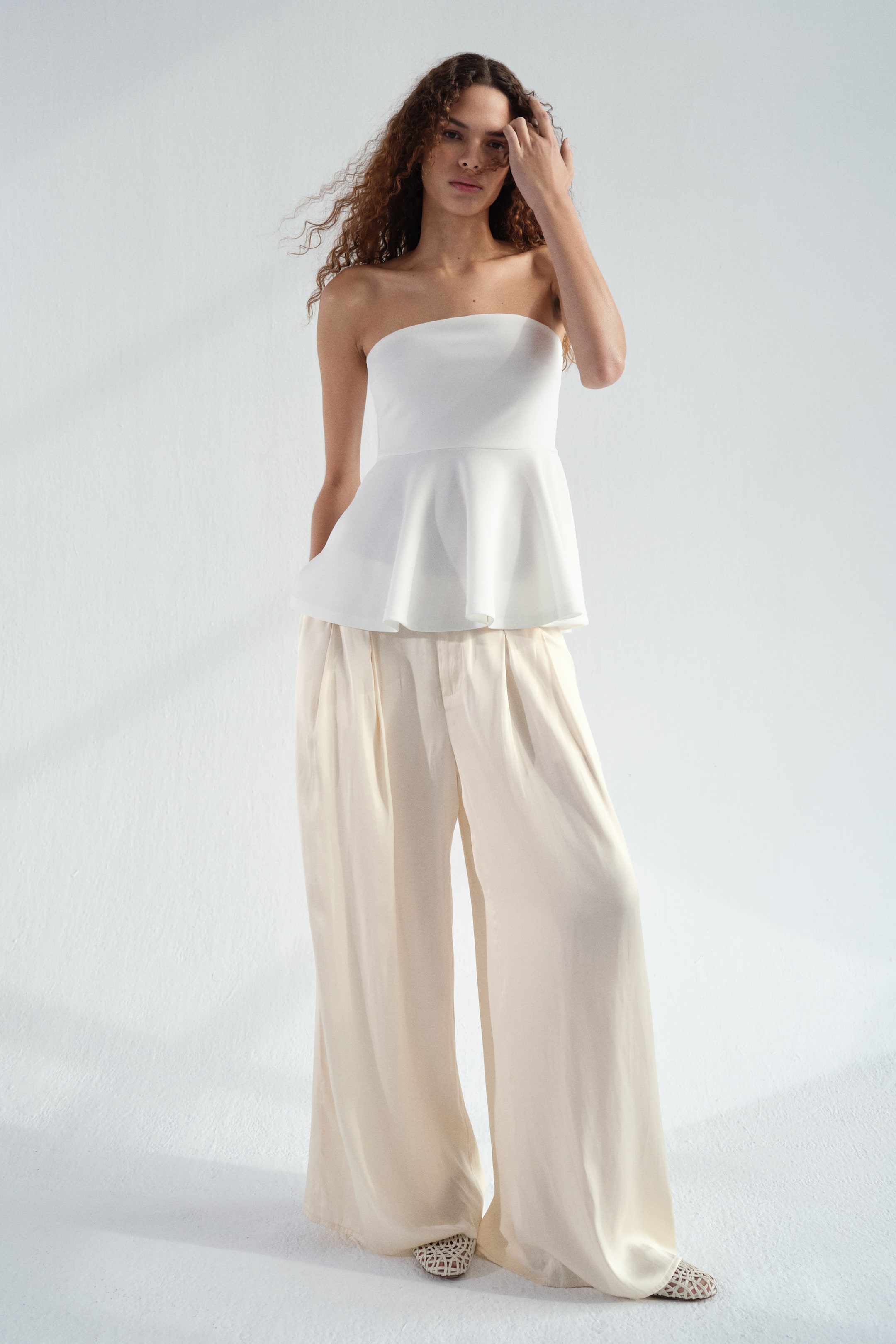 View larger image: The model is showcasing loose-fit satin trousers with an extended waistband and two visible buttons. She stands with one hand casually placed in one of the discreet side pockets. These trousers are designed with wide legs and pleats at the top, perfectly paired with a white strapless top and woven flat shoes.