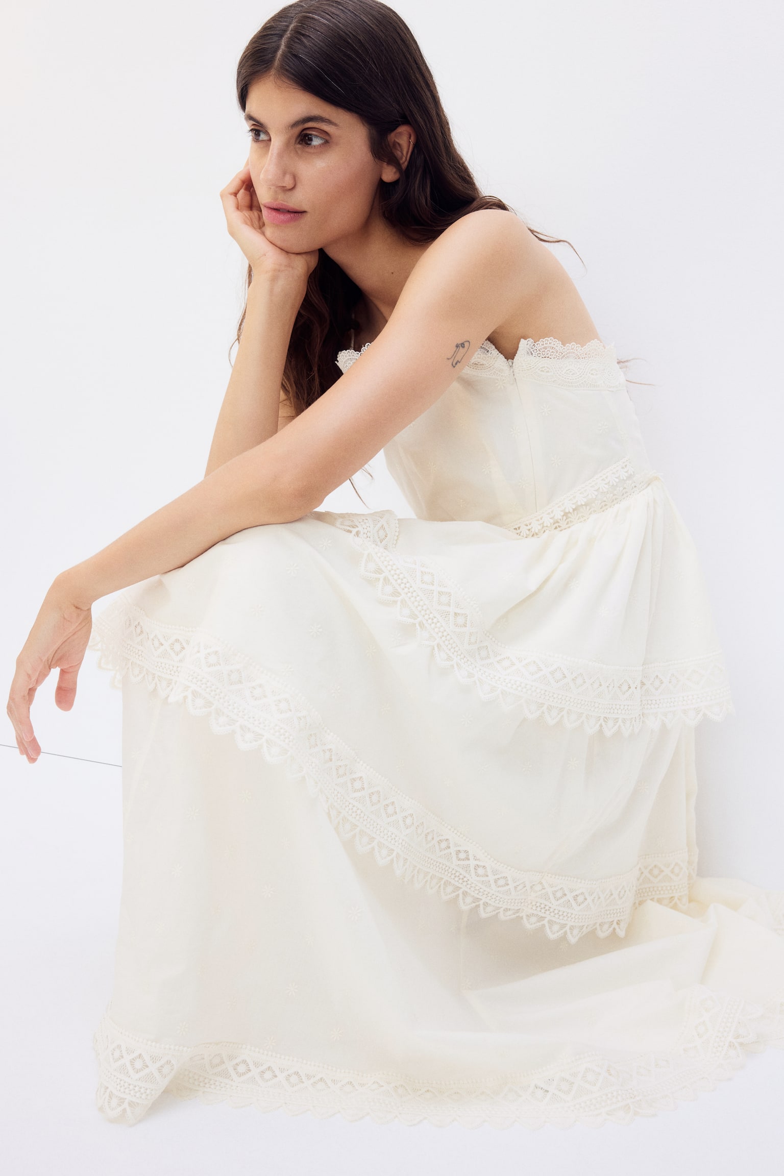 The cream dress is showcased from the front, featuring a fitted bodice and delicate spaghetti straps. It boasts a straight neckline adorned with scalloped lace trim, an embroidered band at the waist, and a full tiered skirt, with each tier highlighting all-over eyelet embroidery and a geometric lace trim with a scalloped edge. - 1