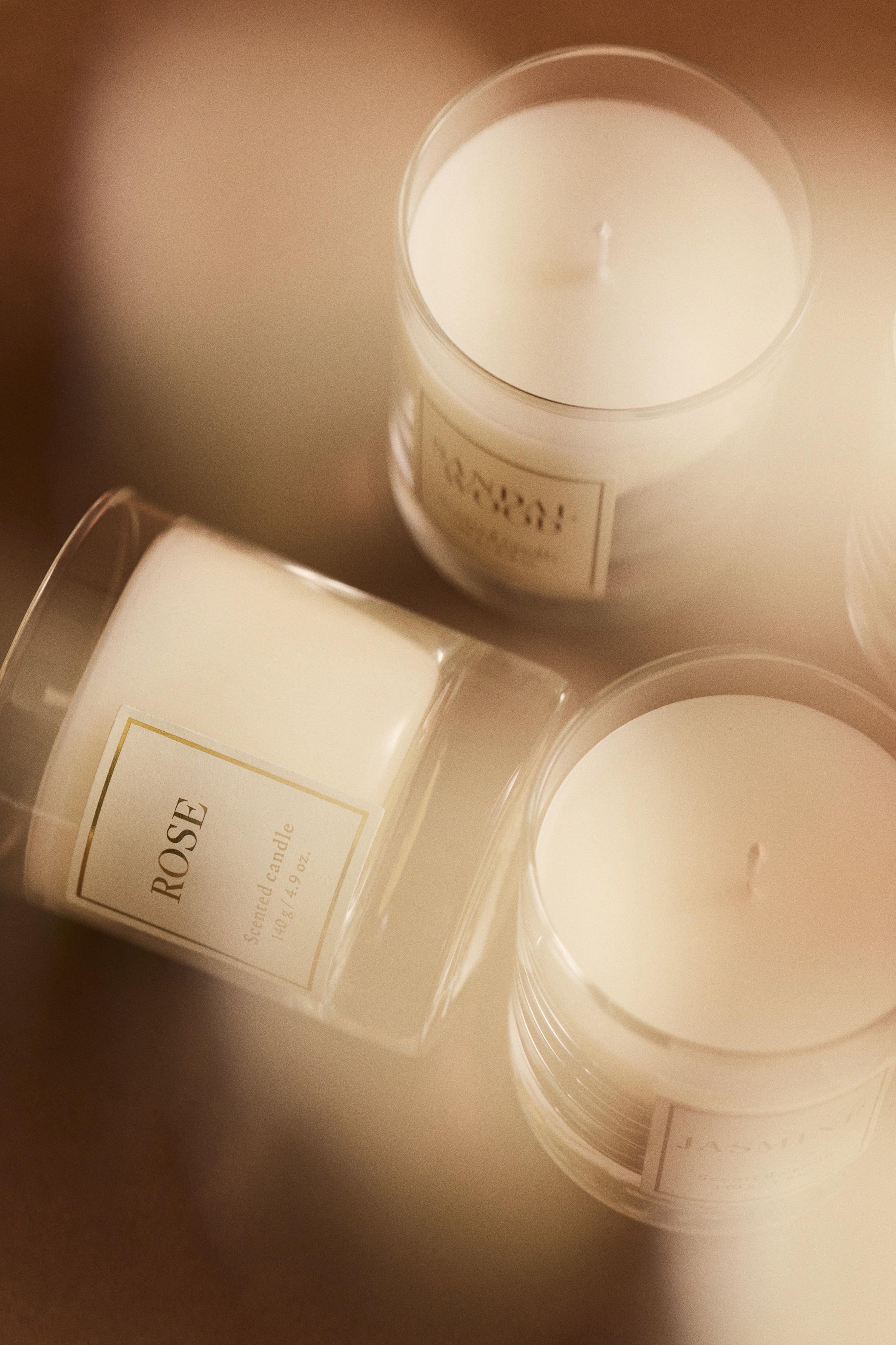 A white scented candle in a clear glass holder with a thick base stands next to its matching burgundy patterned gift box, both facing forwards. Both the candle and the box feature a white rectangular label with a gold border and the word "ROSE" printed in the centre.