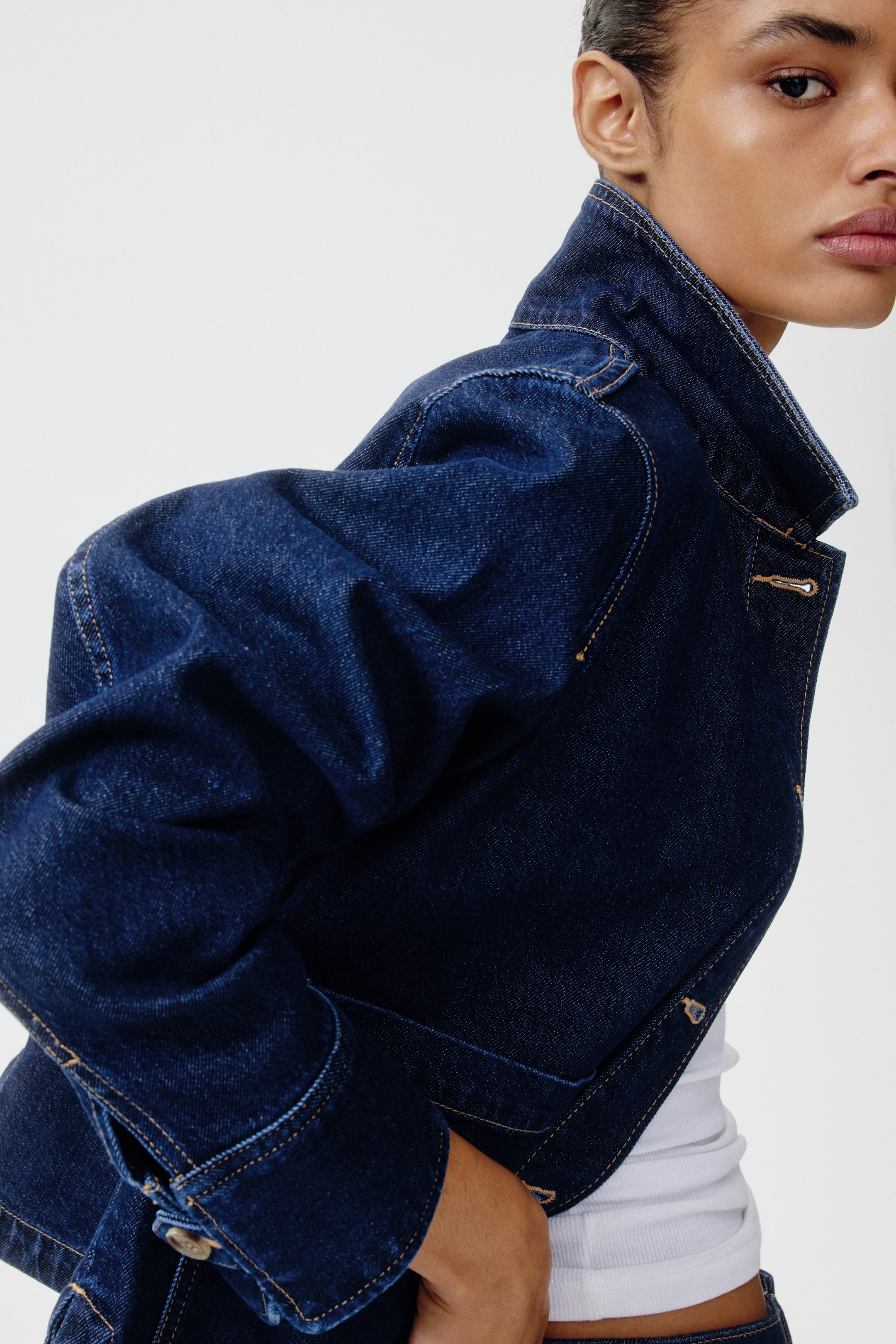 View larger image: A dark blue denim jacket is being presented, angled to the right, prominently featuring contrasting light brown stitching, a standing collar, and buttoned cuffs. A white ribbed top is layering underneath the jacket, visible at the open cuff and bottom edge.