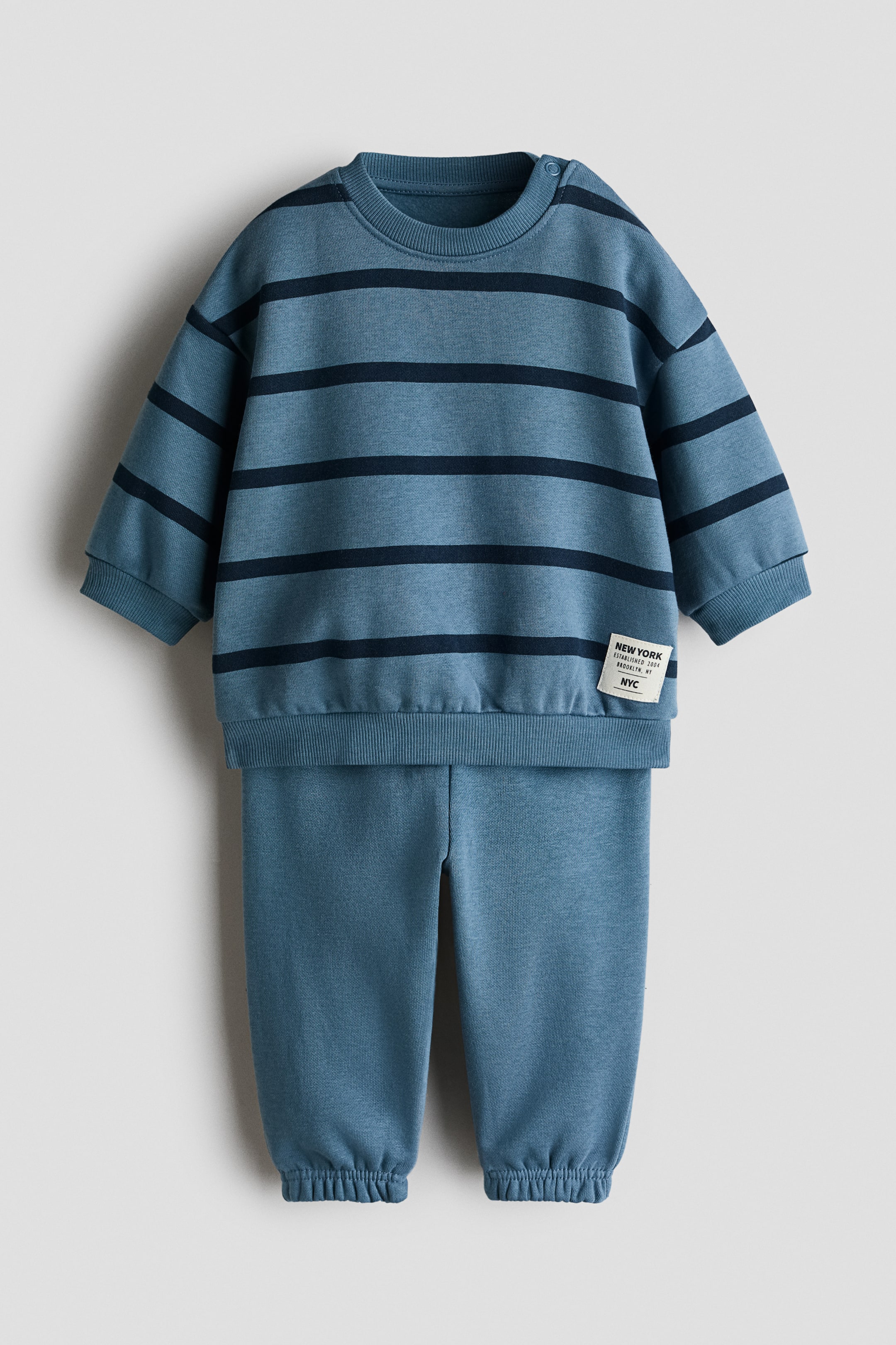 View larger image: Introducing a two-piece sweatshirt set, comprising a blue sweatshirt adorned with dark blue horizontal stripes, a crew neck, shoulder snaps, and ribbed cuffs and hem. The coordinating solid blue sweatpants feature elasticated ankle cuffs.