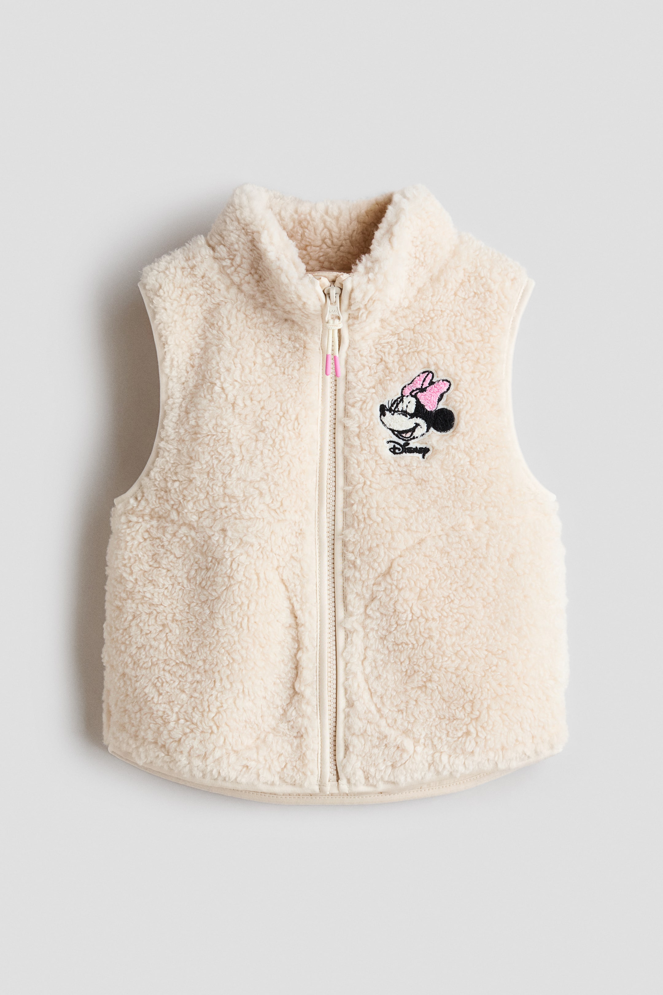Visa större bild: En krämfärgad sherpa‑väst visas framifrån och har en stående krage samt hel dragkedja med en rosa detalj på dragkedjans dragare. En broderad Minnie Mouse‑patch med texten "Disney" syns på vänster bröst.