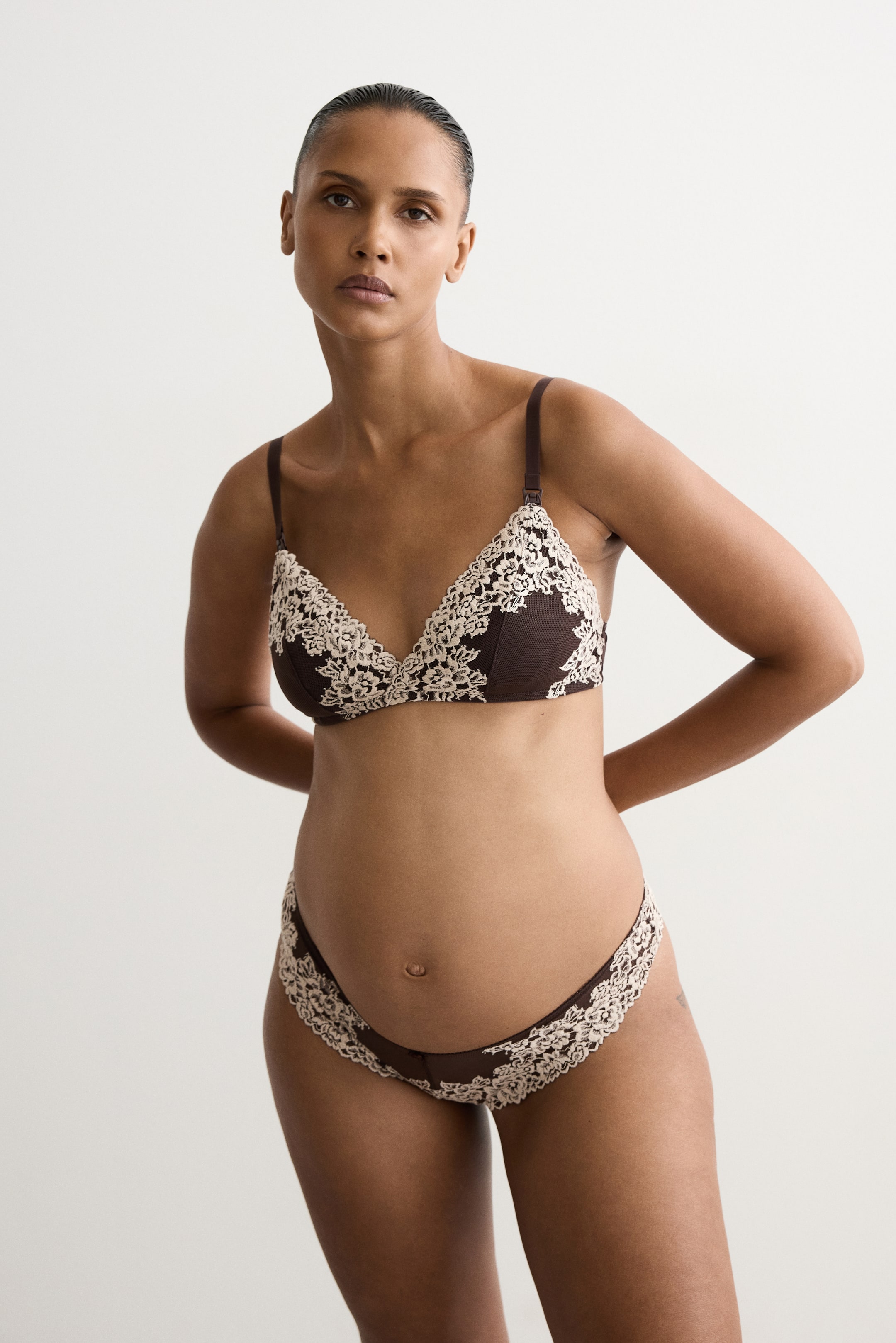 View larger image: A wireless nursing bra in dark brown with cream-coloured lace trim on the cups is being worn by the model, featuring adjustable shoulder straps with nursing clasps. The model is also wearing a matching pair of low-rise briefs with coordinating lace detail.