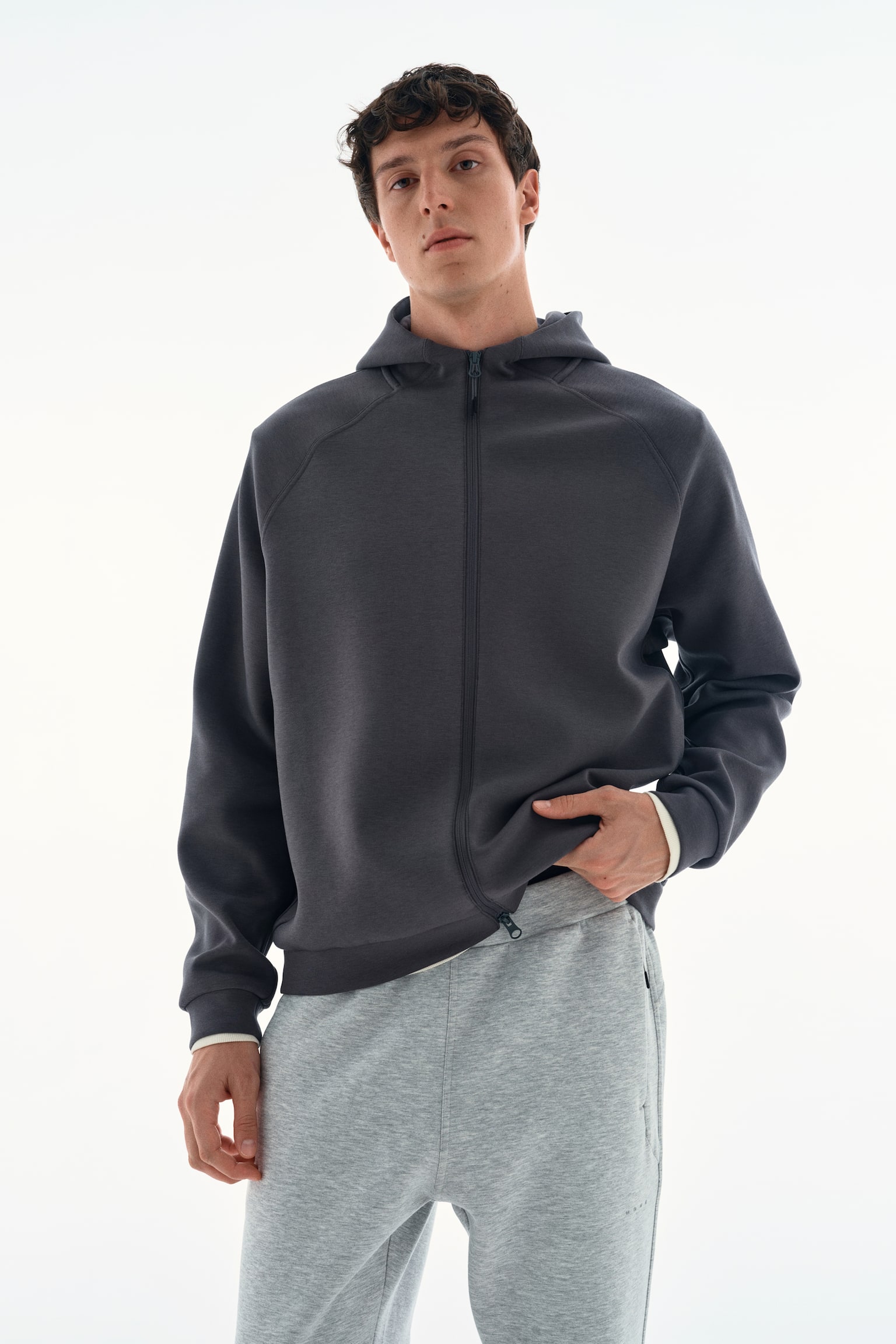 A dark grey zip-up hoodie with raglan sleeves and a full-length front zip is being worn by the model, who is holding the bottom hem slightly to one side. The hoodie is layered over a white top, visible at the cuffs and hem, and is paired with light grey sweatpants. - 1