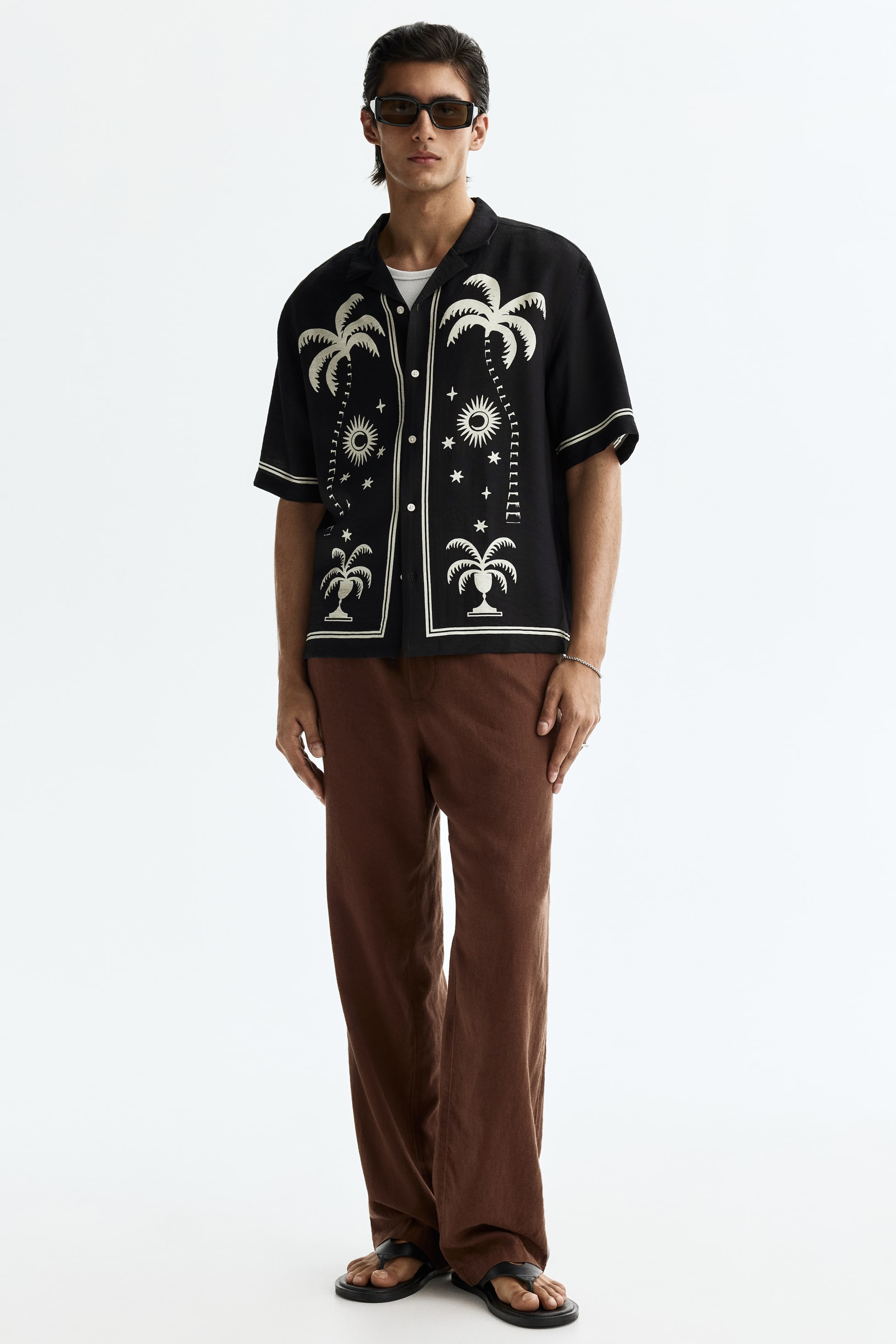 View larger image: A black short-sleeved shirt is facing forward, featuring white embroidered palm trees, suns, moons, and stars. It showcases a camp collar and white piping along the placket, collar, and sleeve hems, and is worn unbuttoned over a white t-shirt.