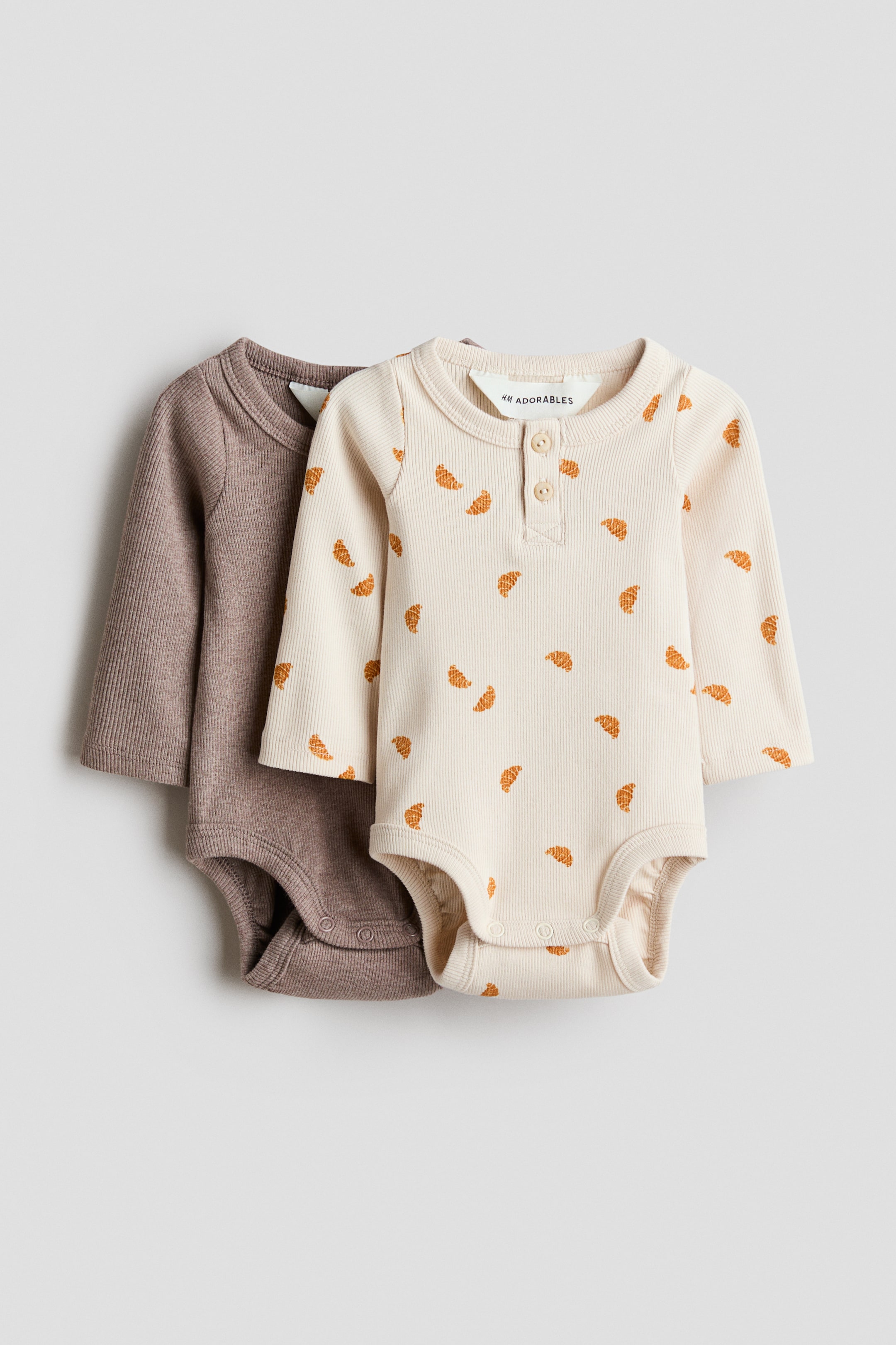 View larger image: Two baby bodysuits are shown: one in solid brown ribbed fabric and the other in light beige ribbed fabric. Both feature an all-over orange croissant pattern, a two-button Henley placket at the rounded neckline, and snap closures at the crotch.