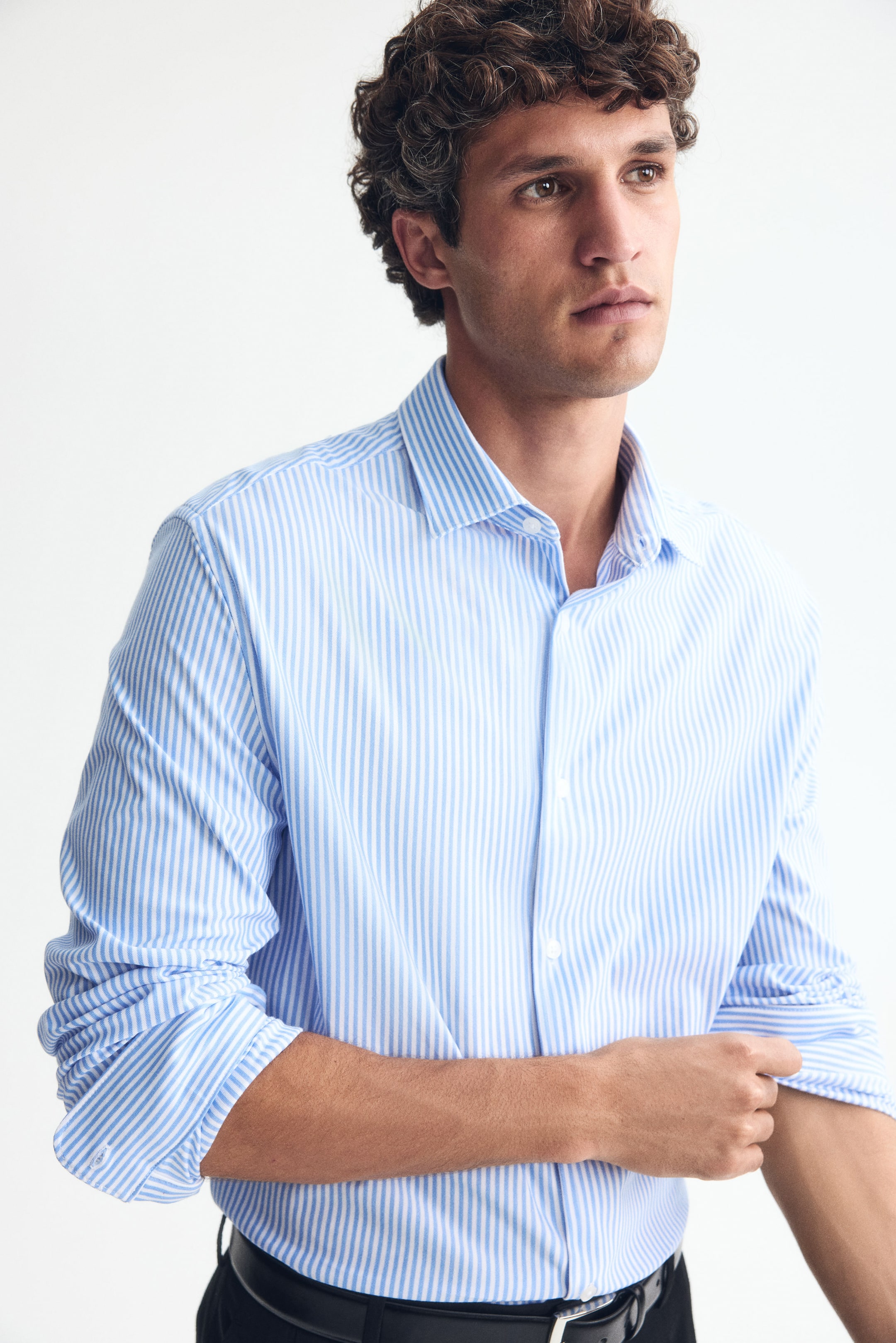 Ampliar la imagen: Una camisa de ajuste slim con rayas azules y blancas, cuello cutaway y frente francés está siendo usada por el modelo. El modelo está arremangándose una de las mangas largas, mientras la camisa está metida dentro de unos pantalones negros.