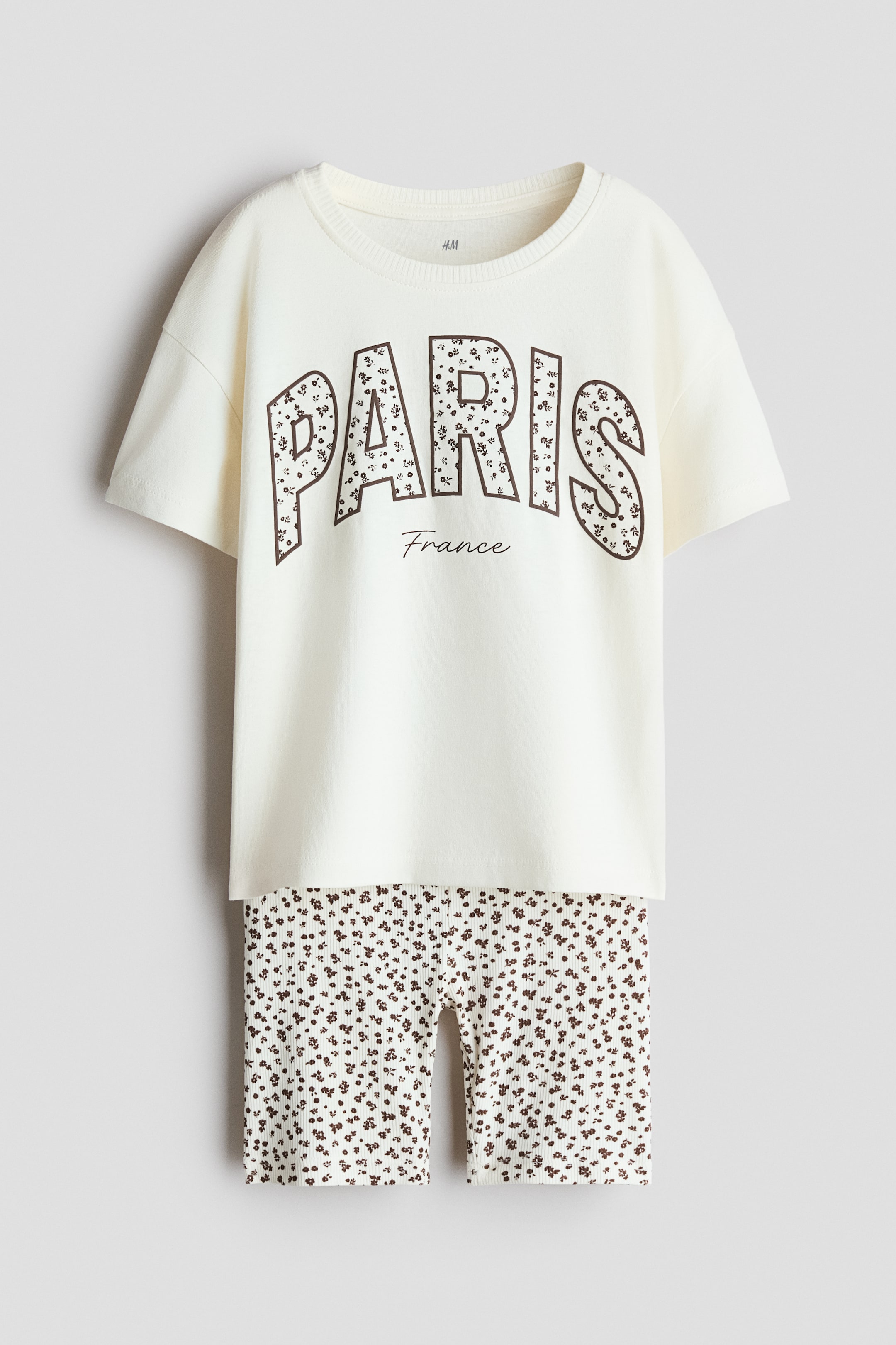 View larger image: A two-piece clothing set is shown from the front, consisting of an off-white crewneck t-shirt and matching shorts. The t-shirt displays the word "PARIS" in large block letters filled with a brown floral print, while the fitted shorts feature the same all-over floral pattern on a ribbed fabric.