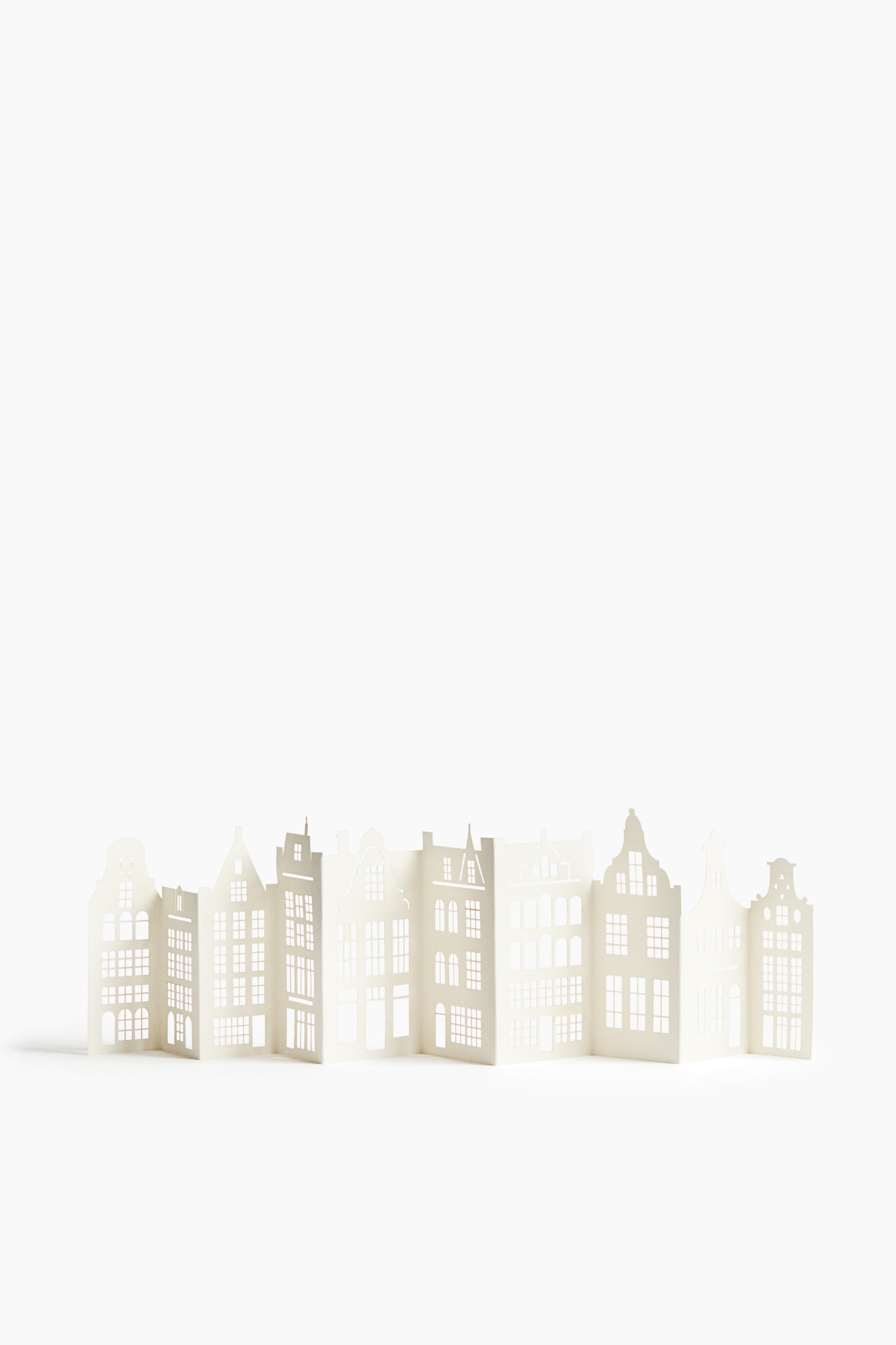 View larger image: A white decorative paper screen stands in a freestanding accordion-fold layout, showcasing a row of traditional building facades. The front-facing product features intricate cutouts representing windows, doors, and various architectural gables.