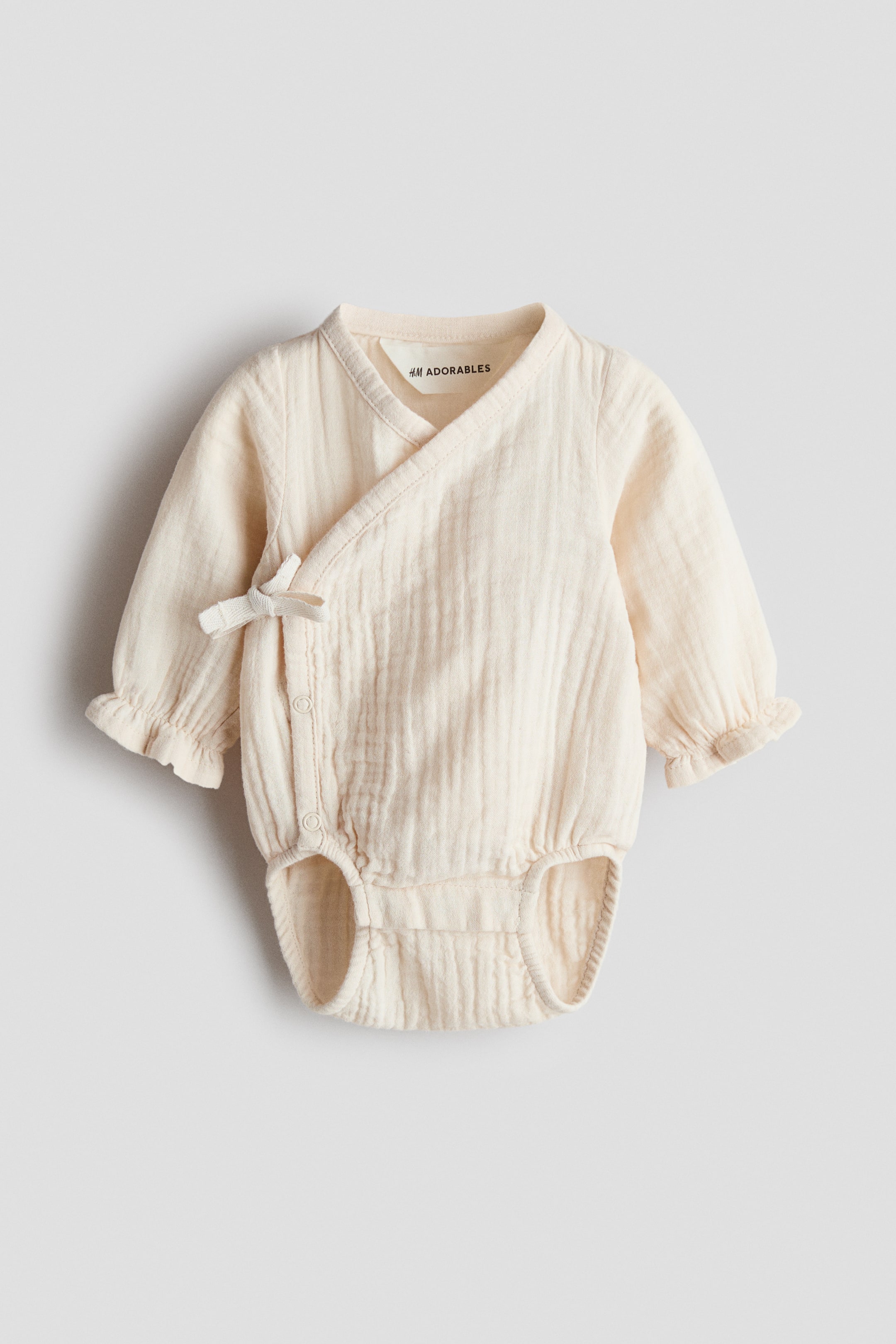 Voir une image plus grande: Une combinaison en mousseline crème de la collection Adorables de H&M est présentée de face, mettant en valeur un design croisé avec un nœud latéral et des boutons-pression visibles, des manches longues avec poignets élastiqués bordés de volants, et des ouvertures de jambes élastiquées.