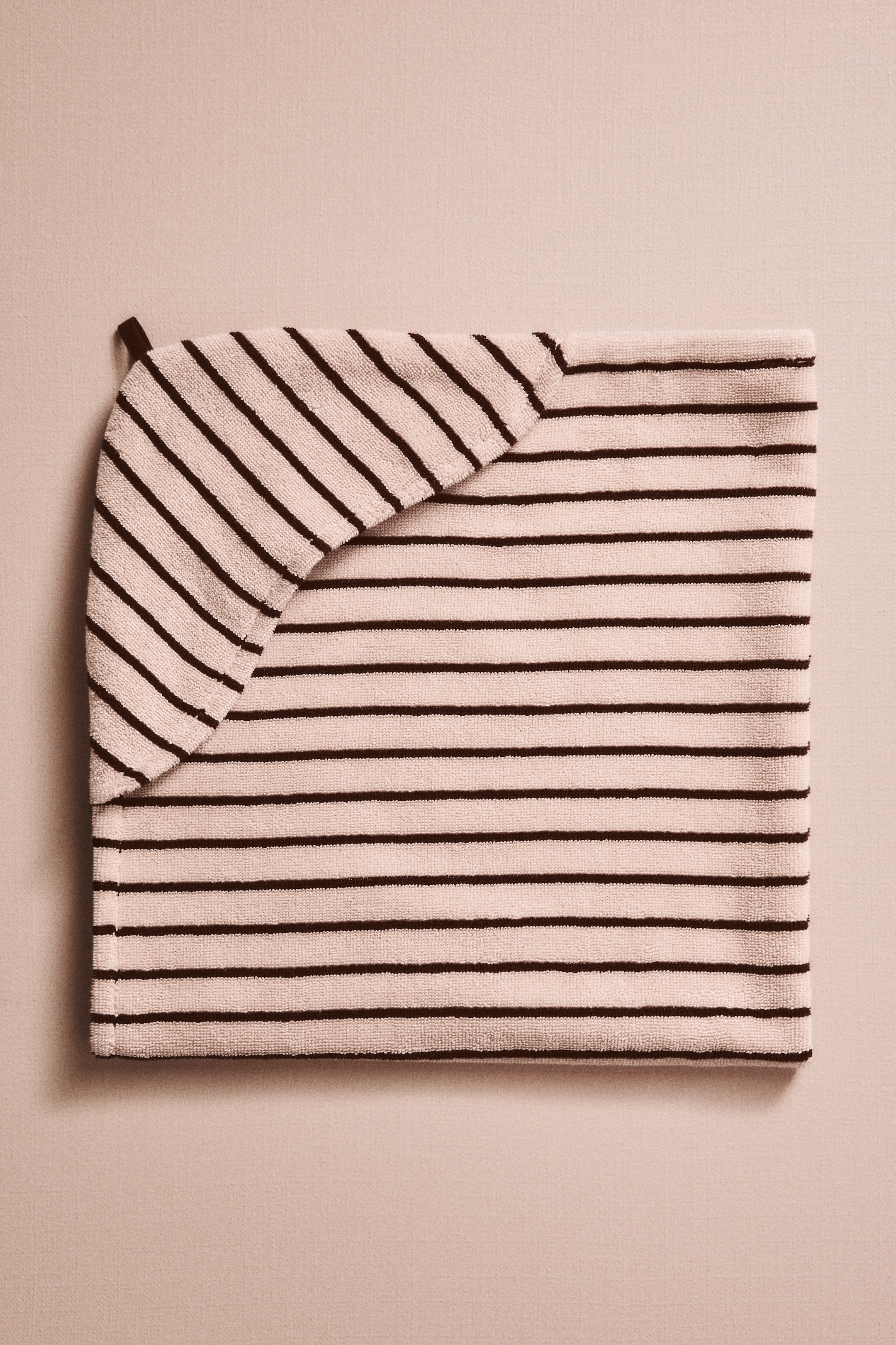 Vezi imaginea mai mare: Un articol textil dreptunghiular este pliat plat, având dungi orizontale închise pe un fundal deschis. O secțiune triunghiulară a articolului se pliază din colțul din stânga sus, afișând dungi diagonale.