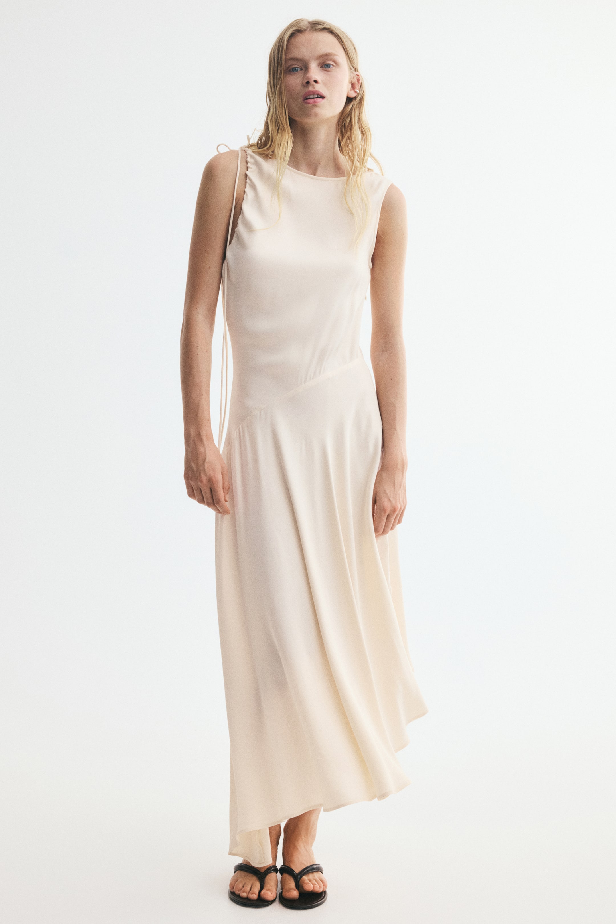 Xem ảnh lớn hơn: Một chiếc váy dài ngang bắp chân, không tay, bằng satin viscose mềm rũ đang được người mẫu mặc, nổi bật với cổ tròn, dây rút buộc quanh một lỗ tay, đường may chéo qua hông và gấu váy bất đối xứng. Người mẫu đang đứng và đi dép sandal màu đen.