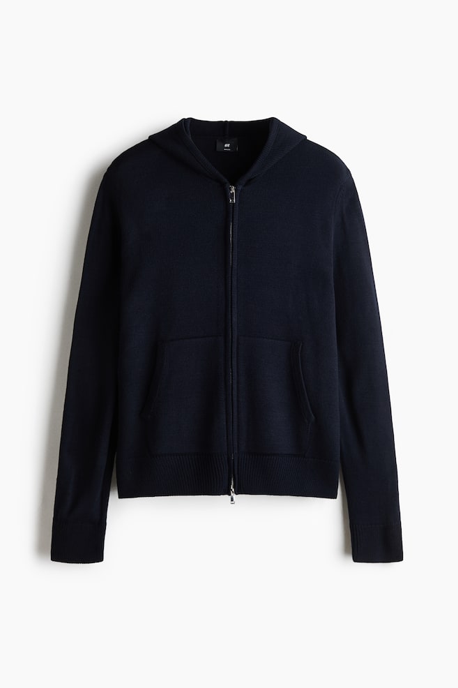 Un cardigan con cappuccio in maglia blu navy scuro è presentato in una vista frontale, caratterizzato da una zip frontale bidirezionale argentata e due tasche applicate frontali. Il design è completato da maniche lunghe con polsini a coste e un bordo a coste coordinato. - 1