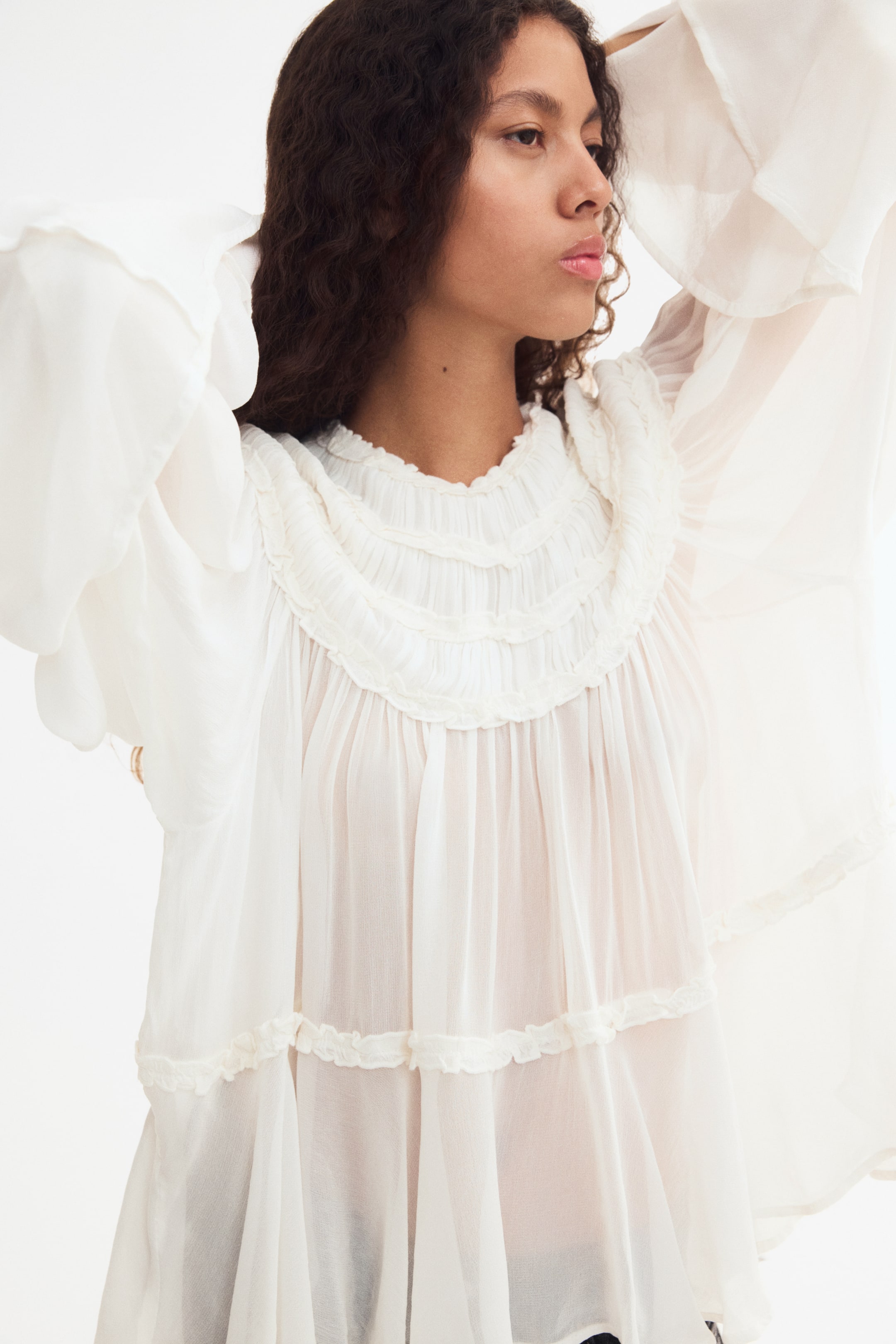 Größeres Bild ansehen: Das Model trägt eine locker geschnittene, halbtransparente weiße Bluse und hält die Arme erhoben, um die ausgestellten Ärmel hervorzuheben. Die Bluse verfügt über einen runden Ausschnitt mit einer geschwungenen, gerafften Passe und Rüschenbesätzen sowie einen horizontalen Rüschenbesatz über dem Saum.