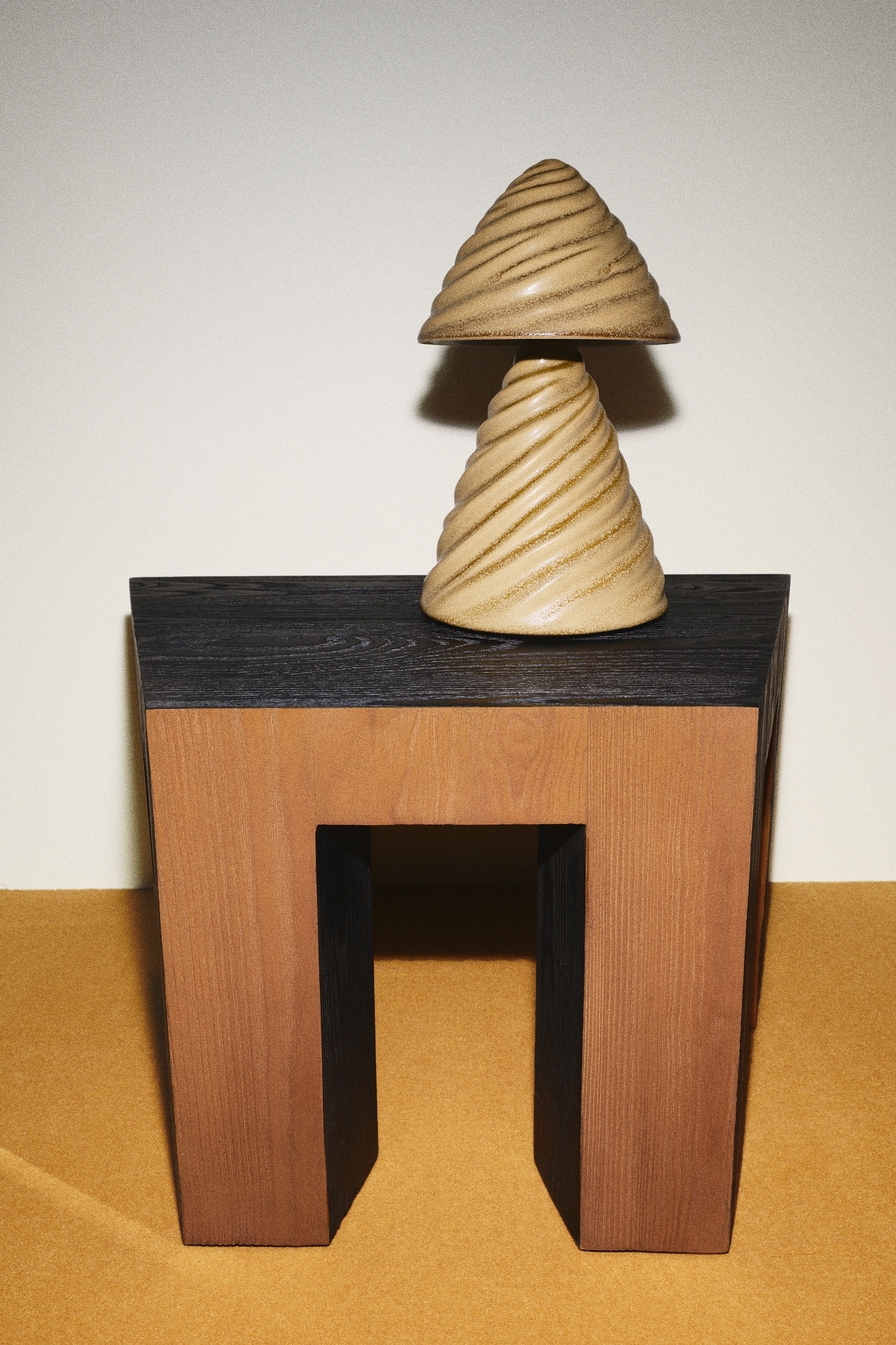 Une lampe texturée de couleur beige clair se tient debout, composée de deux sections coniques avec des crêtes en spirale proéminentes. La section supérieure sert d'abat-jour, reposant au-dessus de la base, et un cordon d'alimentation noir s'étend depuis le bas de la lampe.