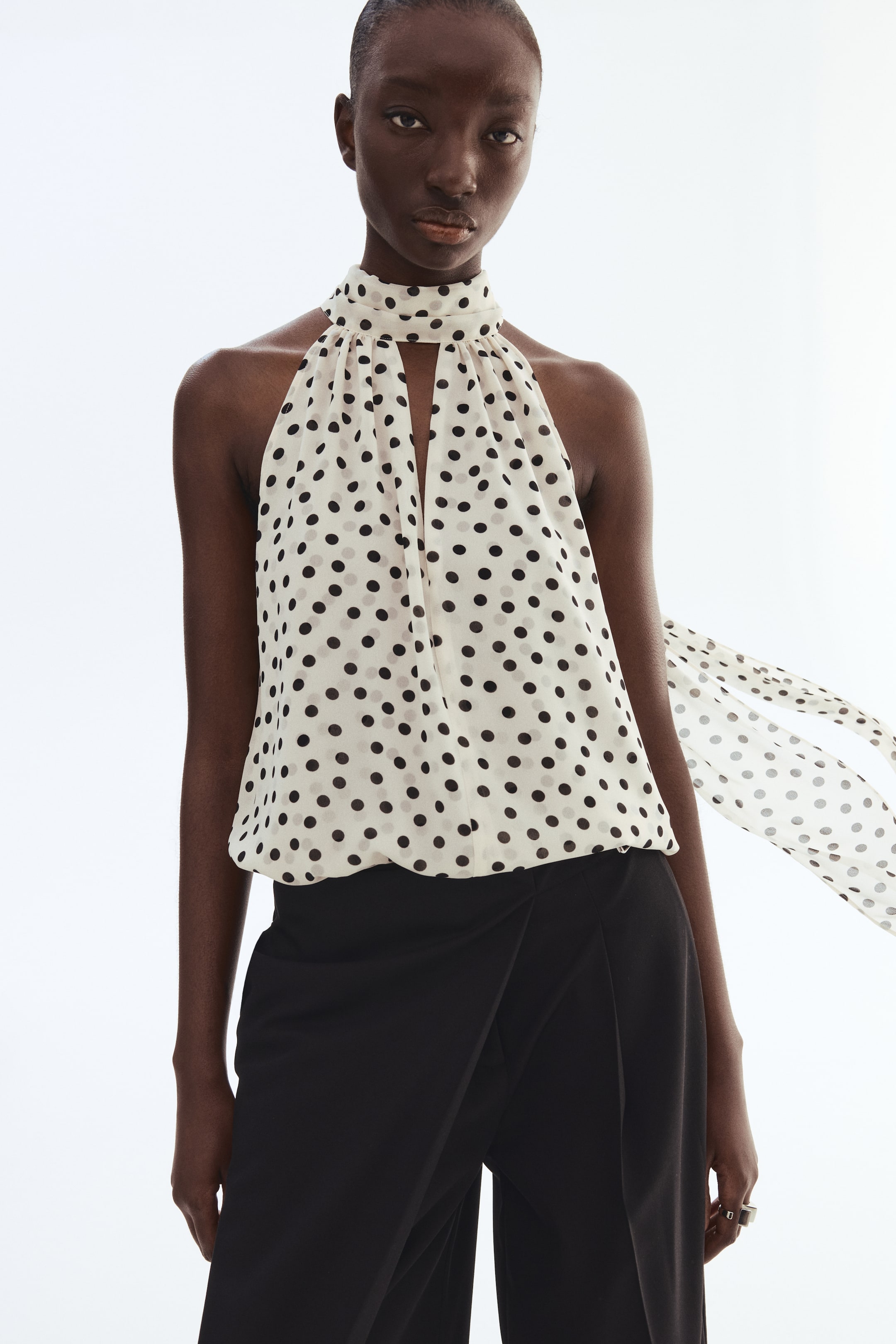 View larger image: The model is donning a loose-fit, sheer top adorned with black polka dots against an off-white backdrop. This piece is highlighted by a scarf collar that creates a deep V-shaped neckline at the front, with one end of the collar elegantly draping down.