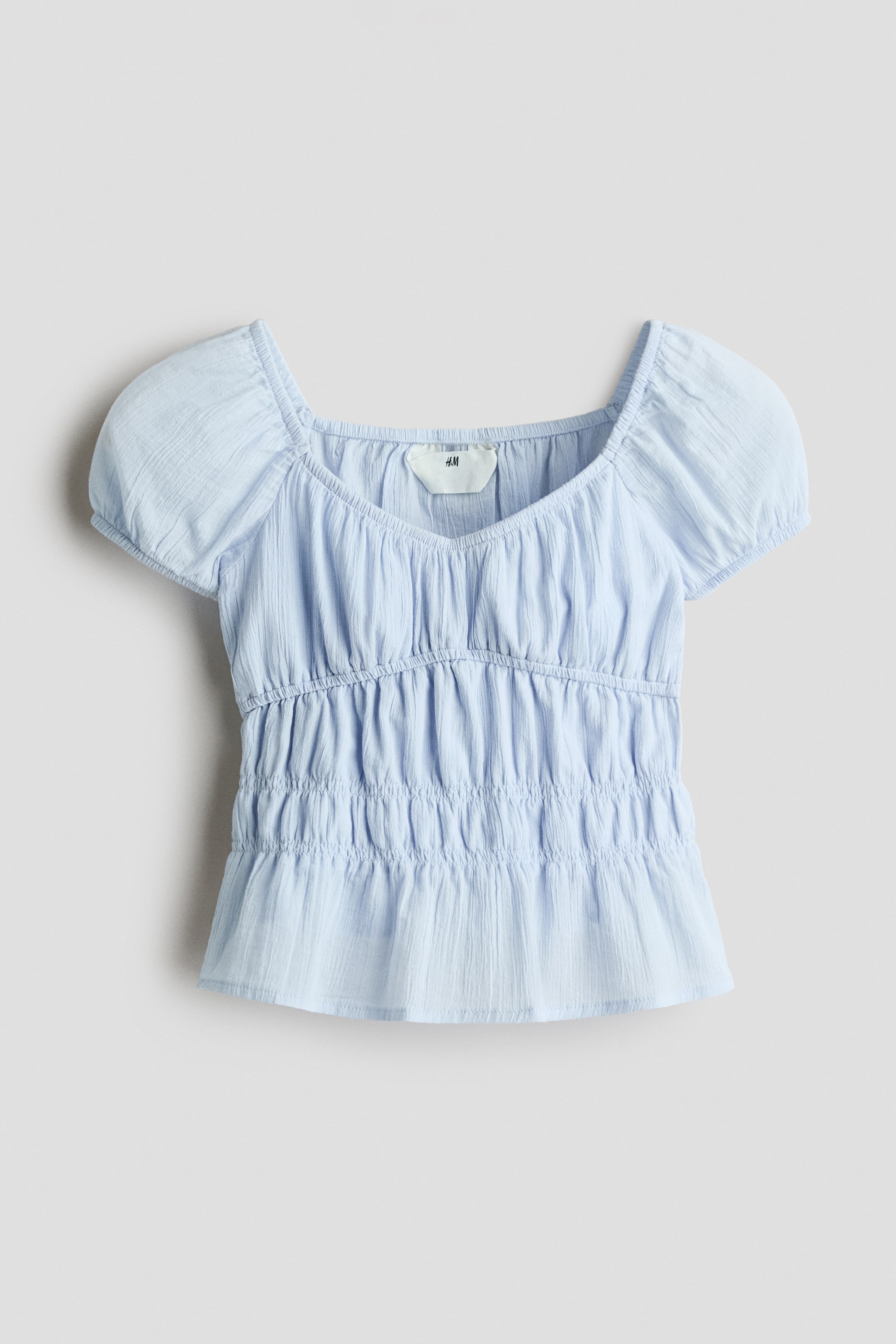 View larger image: A light blue top is shown from the front. It features a square neckline, short puff sleeves, and extensive shirring across the bust and midsection, finishing with a slightly flared hem.