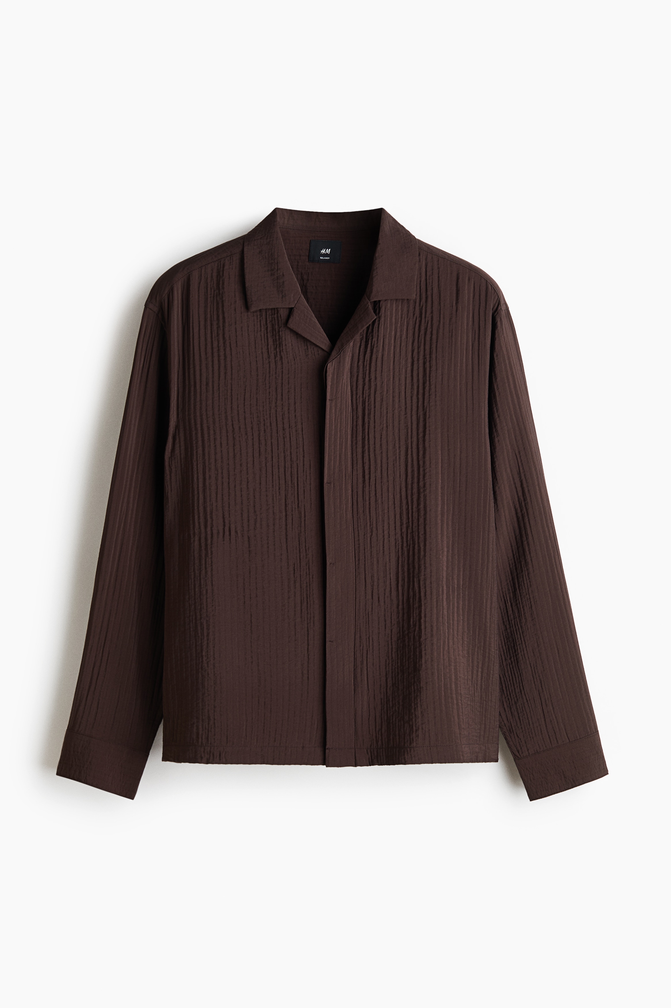 A dark brown, textured-weave shirt with a resort collar and long sleeves is worn unbuttoned by the model. The relaxed-fit shirt is layered over a beige knitted top and paired with dark drawstring trousers.
