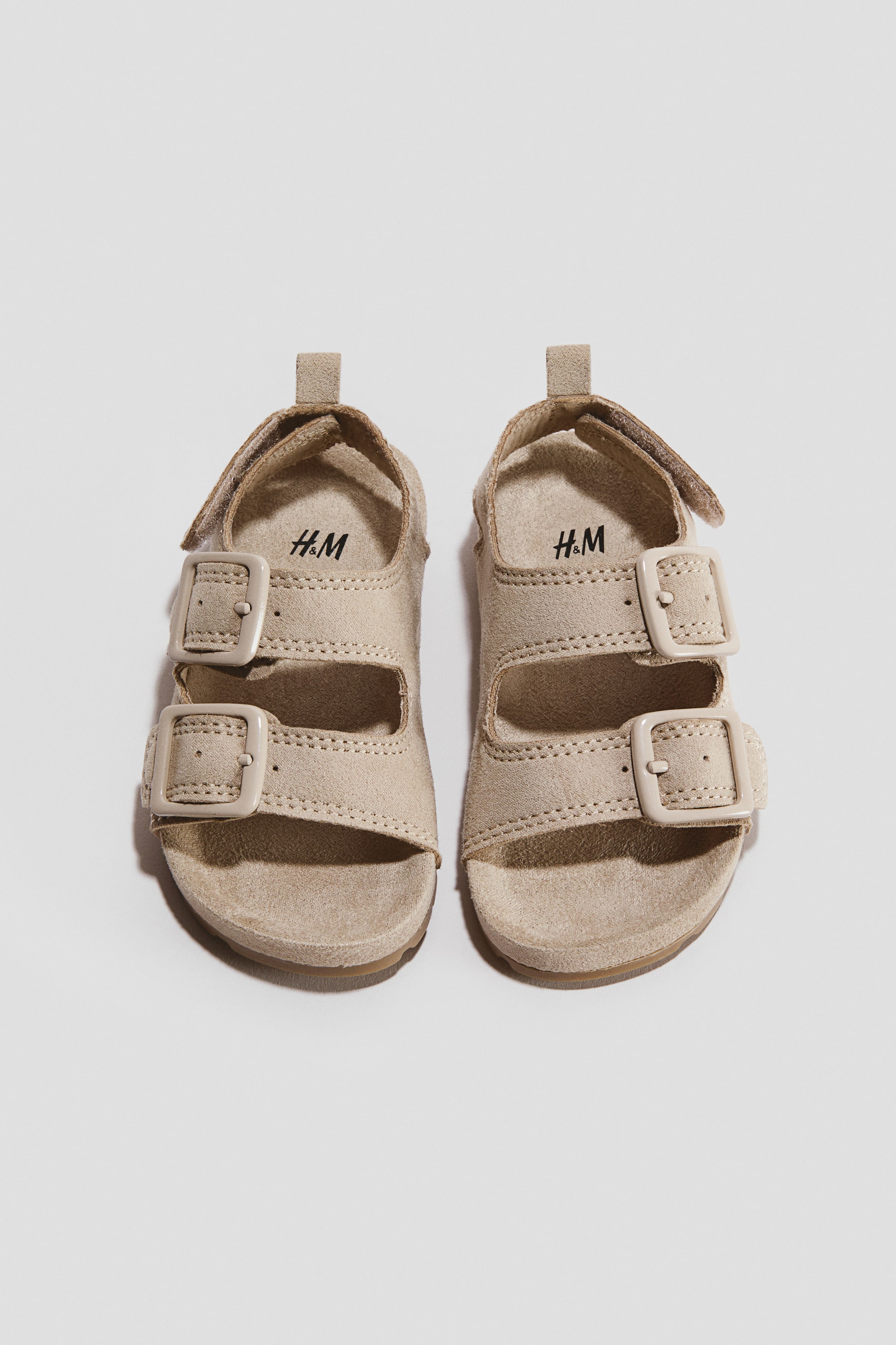 View larger image: Beige suede-textured sandals are shown from a front overhead angle, featuring two adjustable foot straps with colour-matched buckles. The design includes moulded insoles, an ankle strap with a hook and loop fastening, and a small pull loop at the back heel.