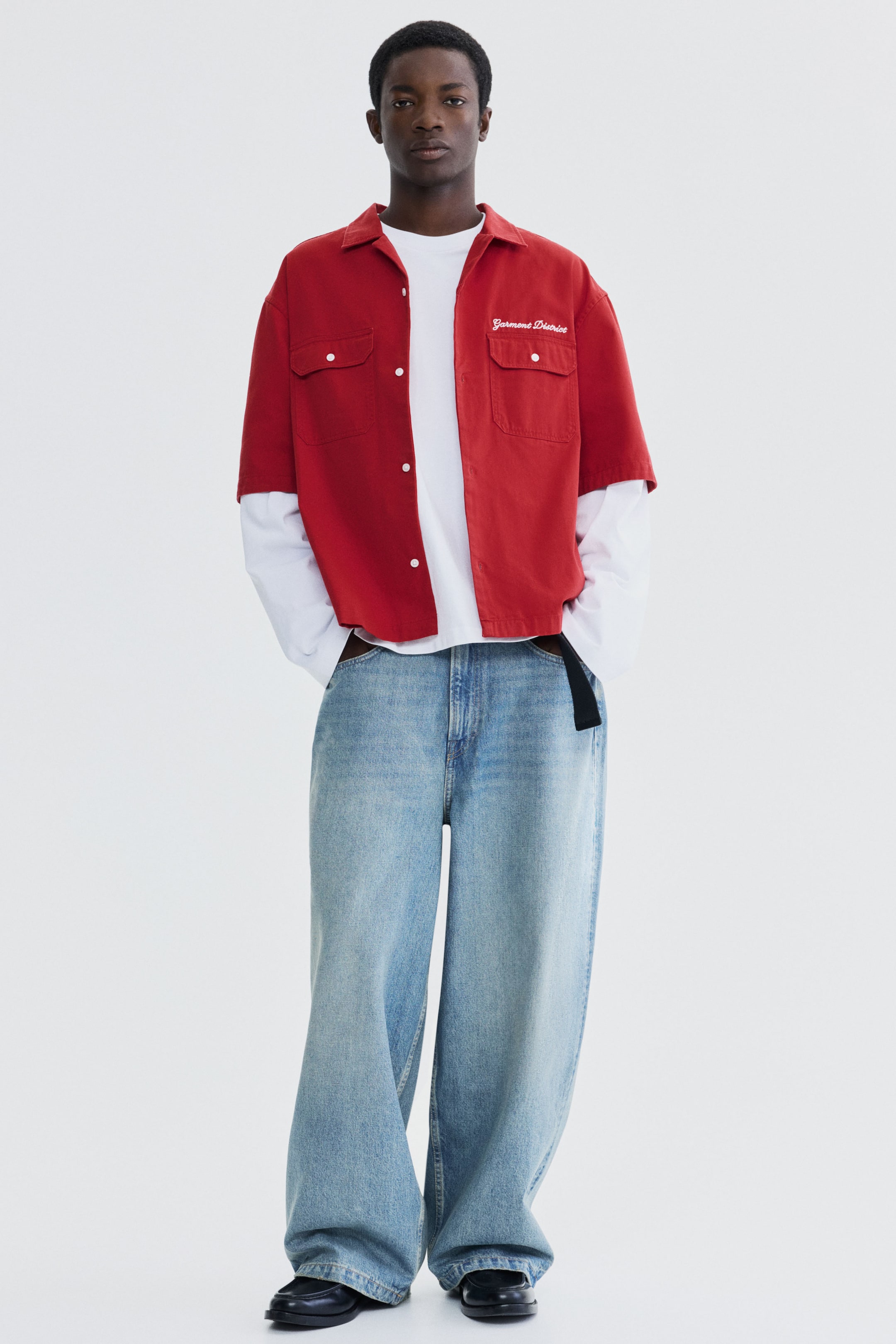 View larger image: A red short-sleeve collared shirt is layered over a white long-sleeve shirt, featuring two buttoned chest pockets and white script embroidery on the right chest. Wide-leg light blue jeans are paired with a partially visible black belt, and black loafer-style shoes complete the outfit.