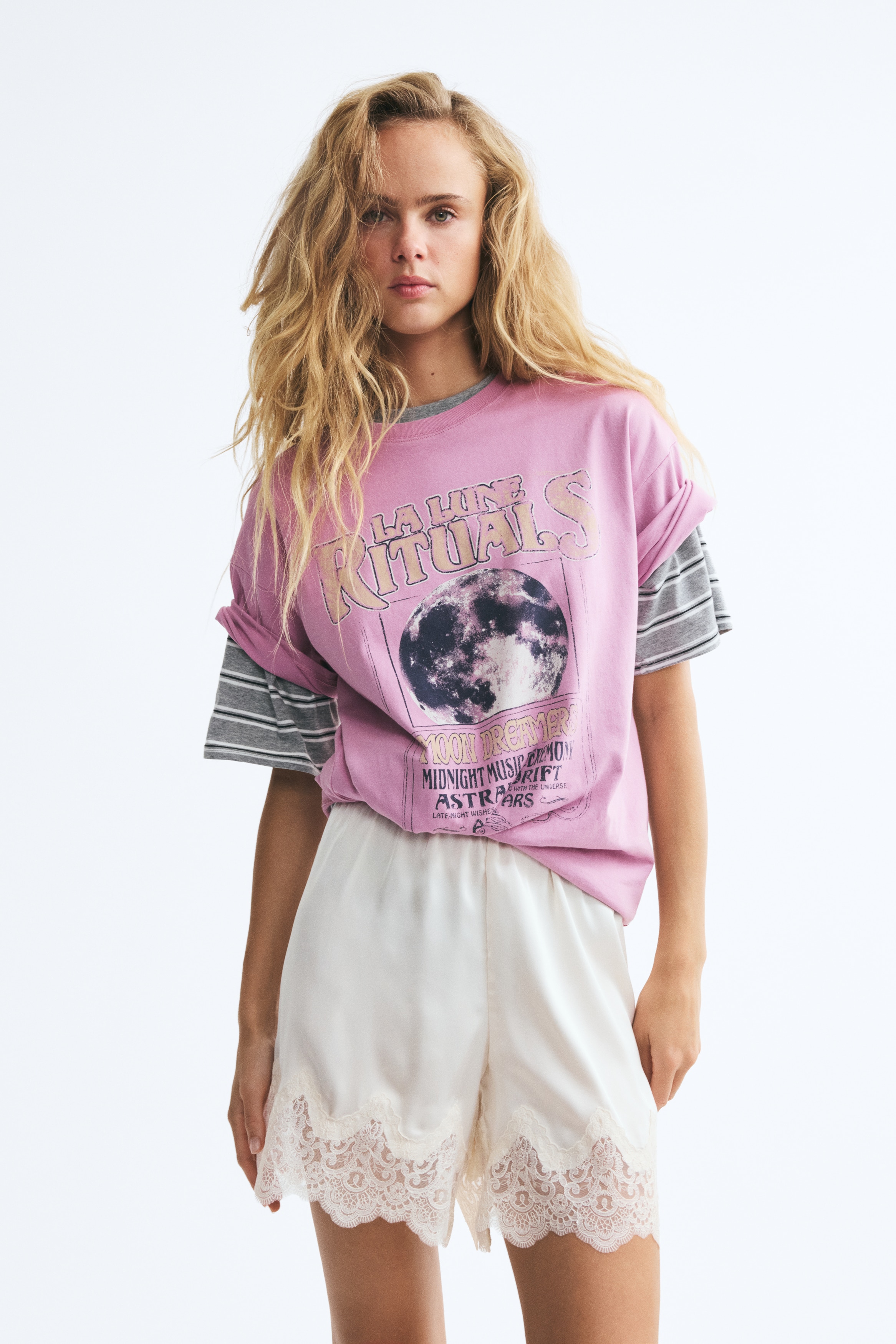 Má»t chiếc áo thun oversized mà u há»ng nhạt ÄÆ°á»£c chụp từ phÃa trưá»c, vá»i cá» tròn, vai trá»
và phom dáng thoải mái. Mặt trưá»c in há»a tiết graphic hiá»u ứng distressed, gá»m hình minh há»a mặt trÄng tròn á» trung tâm, ÄÆ°á»£c bao quanh bá»i dòng chữ cách Äiá»u phong cách vintage "LA LUNE RITUALS" và "MOON DREAMERS".